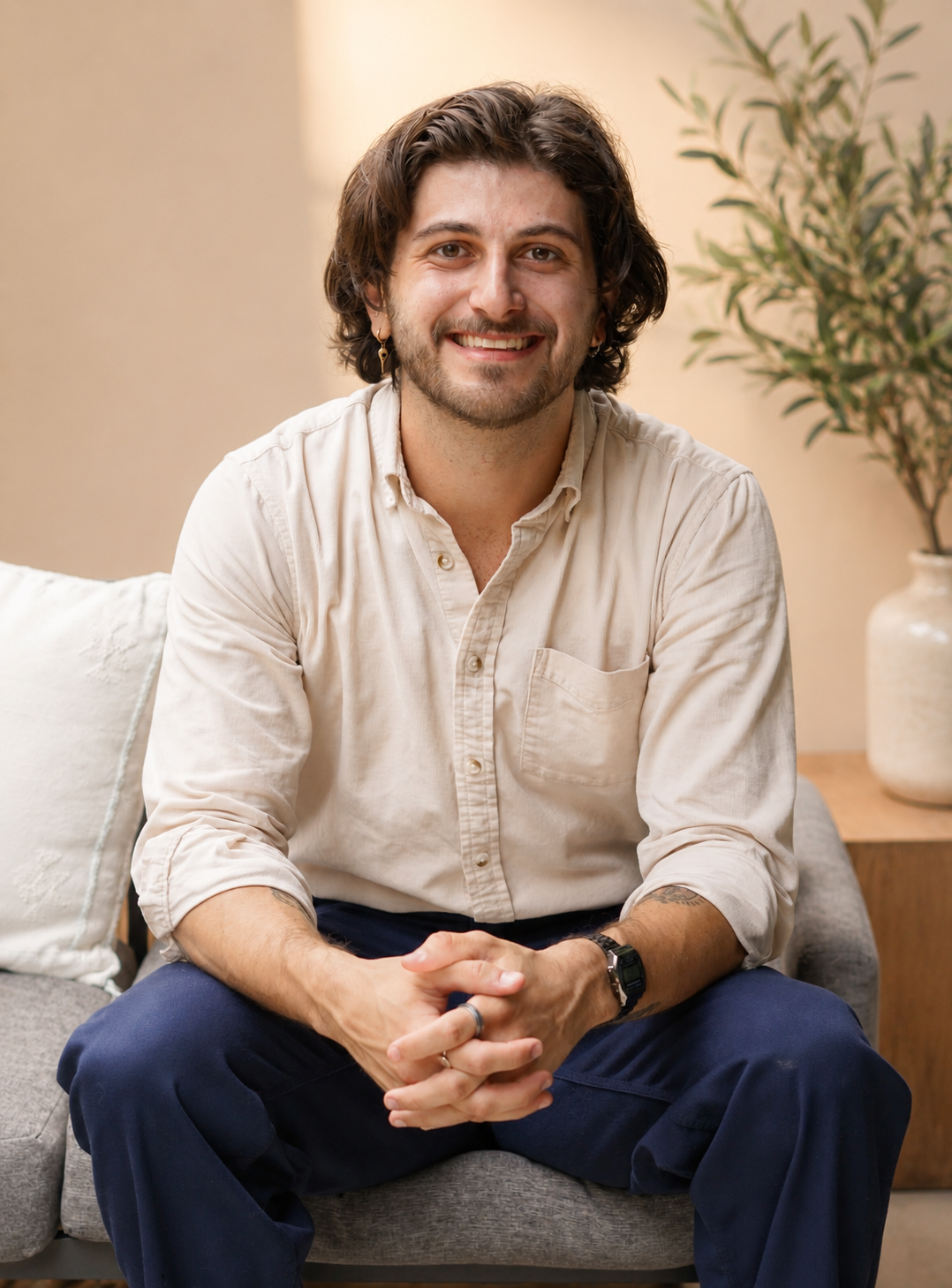 A man smiling, sitting on a gray couch in a cozy living room, with a white pillow and a potted plant in the background.