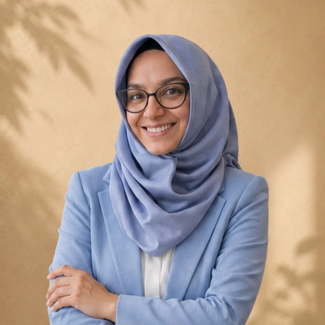 A woman wearing glasses and a blue hijab, smiling, with a light beige background and blurred foliage.