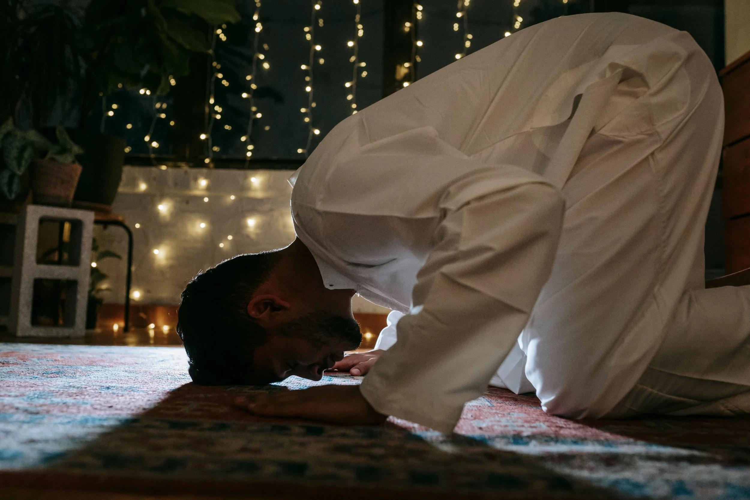 A person mid-prayer, head bowed, expression tense rather than peaceful, soft indoor lighting