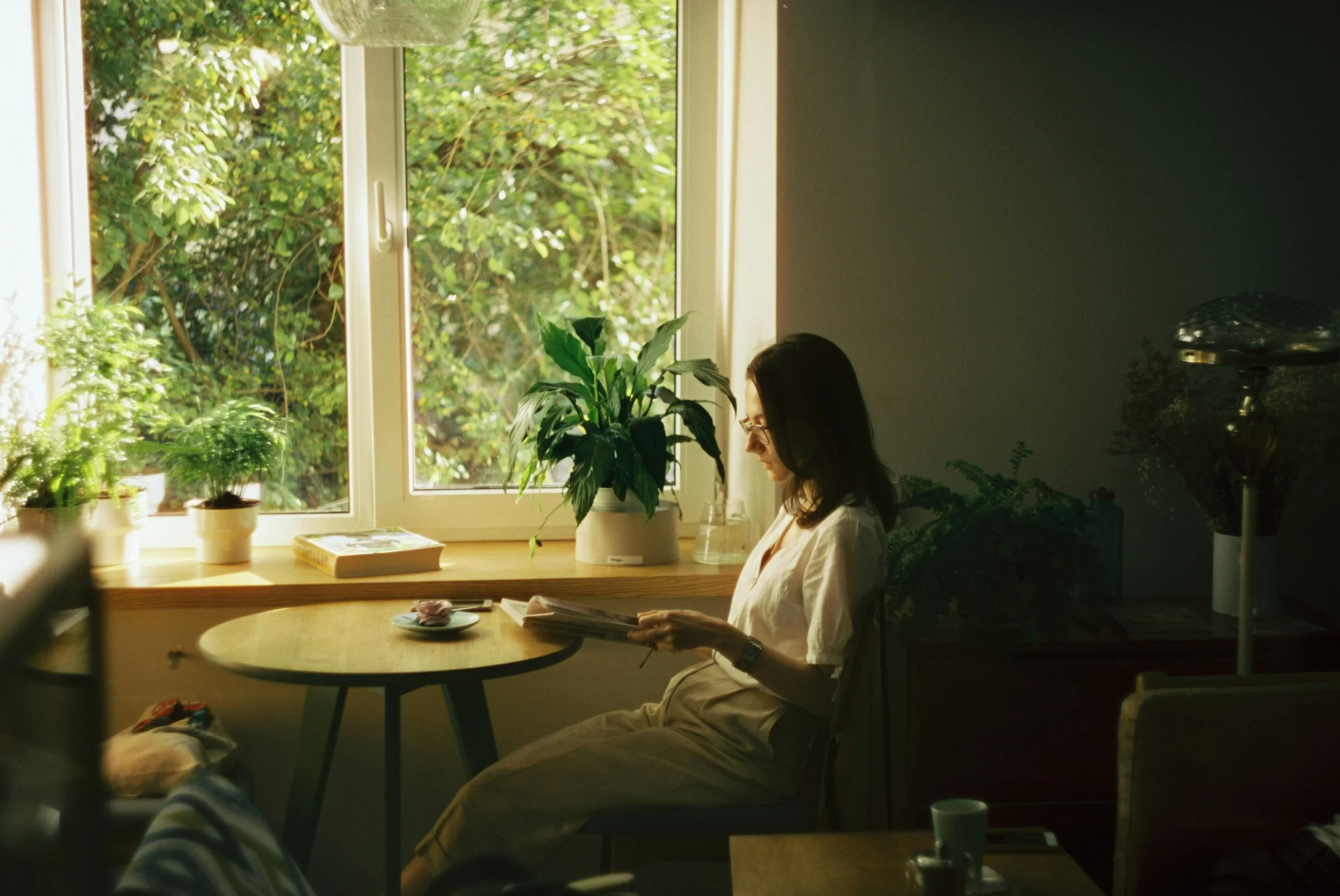 A woman sitting at a round table reading a book or magazine in a cozy room with large windows showing greenery outside, surrounded by potted plants.