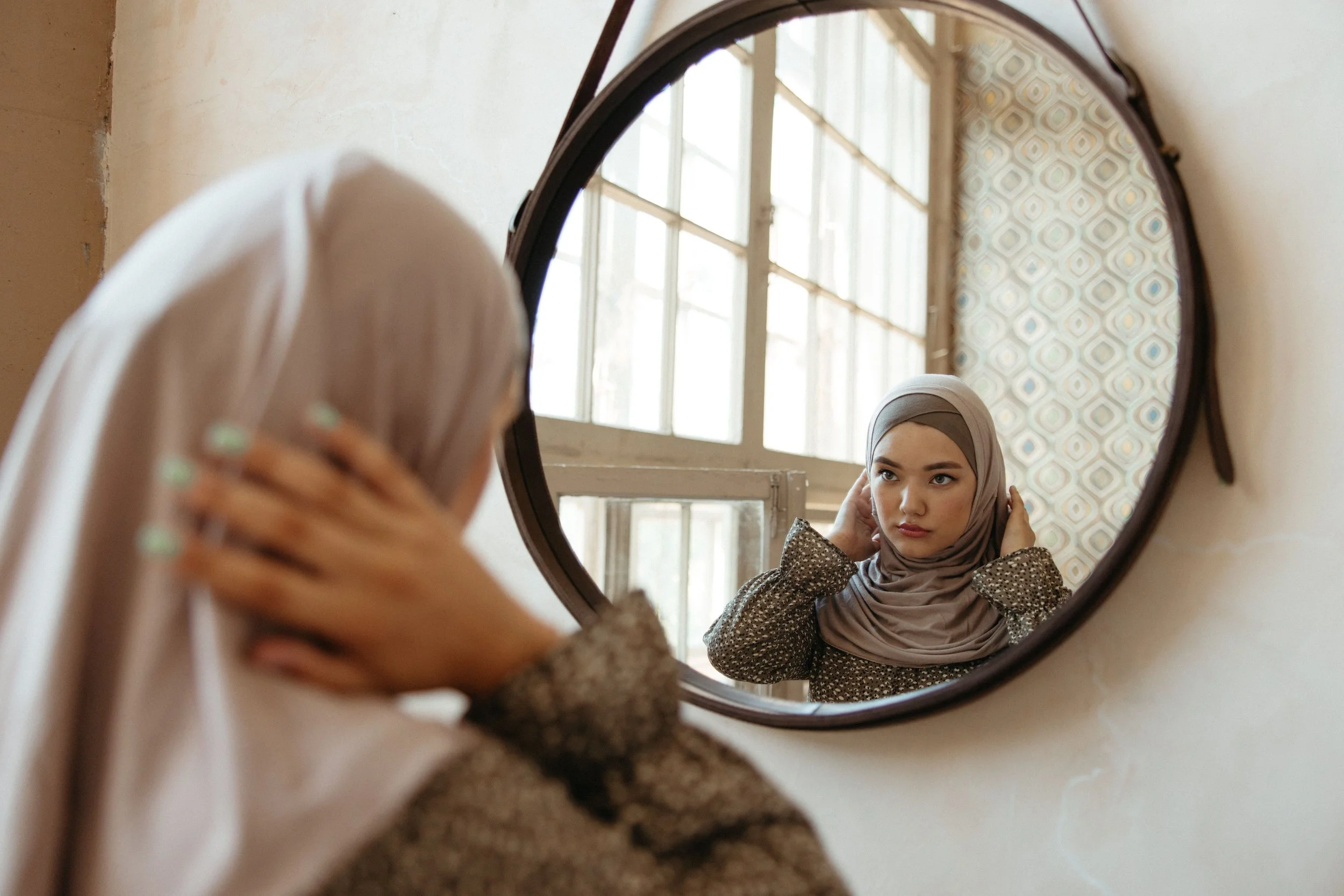 A Muslim woman standing in front of a mirror, expression contemplative, soft and gentle lighting, no focus on body shape