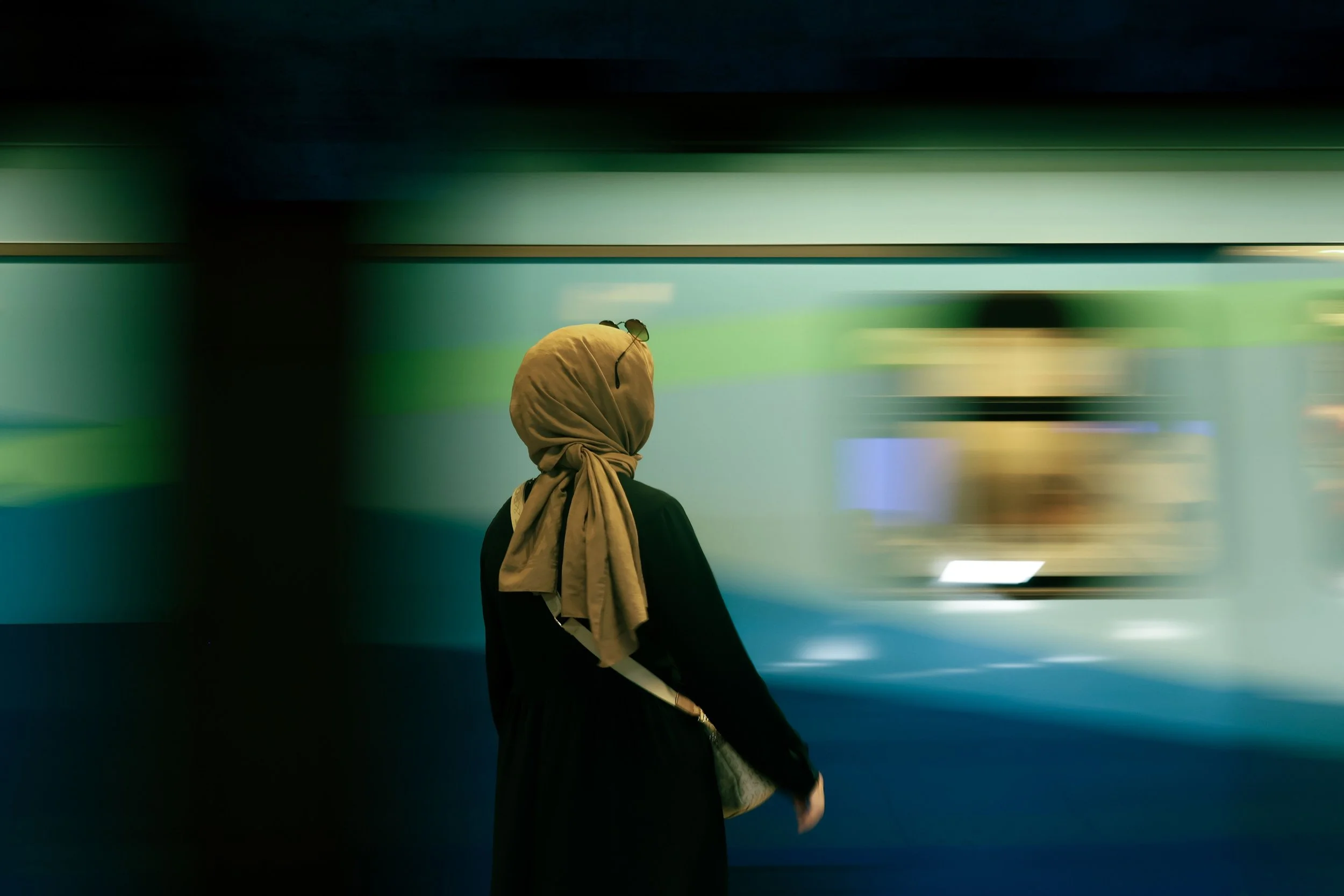 A woman with a beige headscarf and glasses on her head is standing and looking to the left in a dimly lit room with colorful blurred lights in the background.