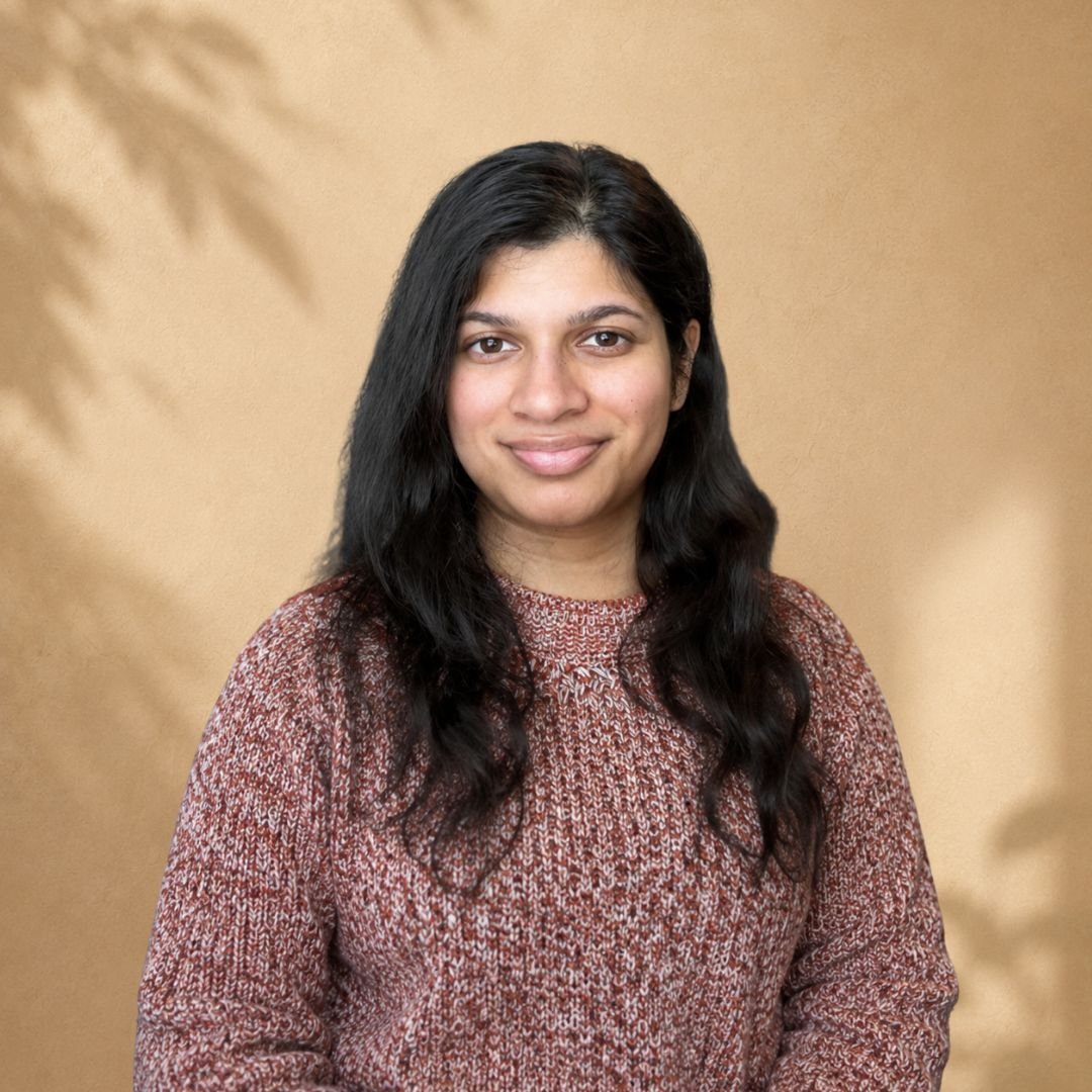 Portrait of a young woman with wavy black hair wearing a patterned long-sleeve shirt, standing against a beige background with blurred shadows.