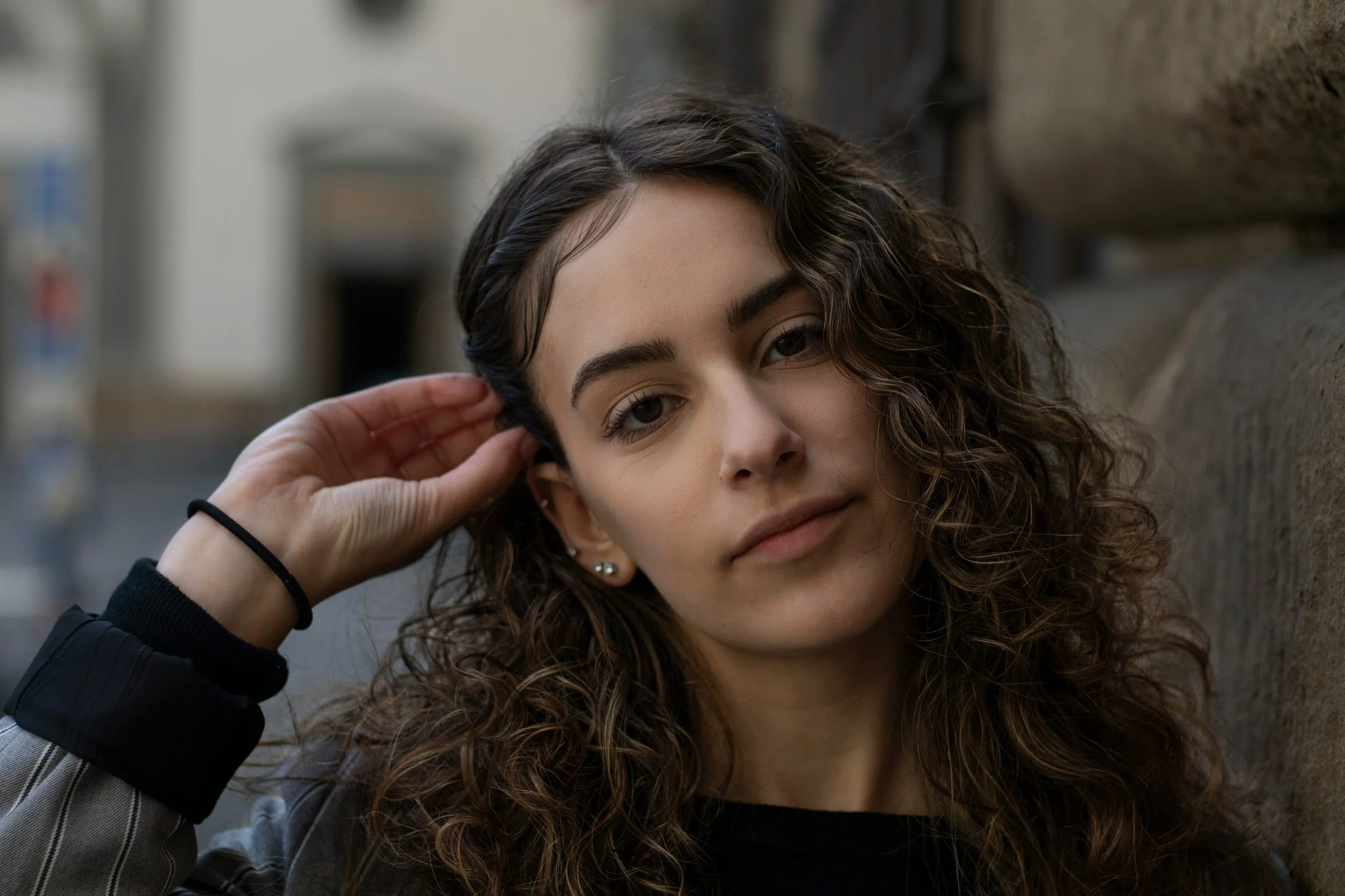 A young woman with wavy brown hair leaning against a brick wall, gently touching her ear with her right hand, and looking at the camera with a relaxed expression.