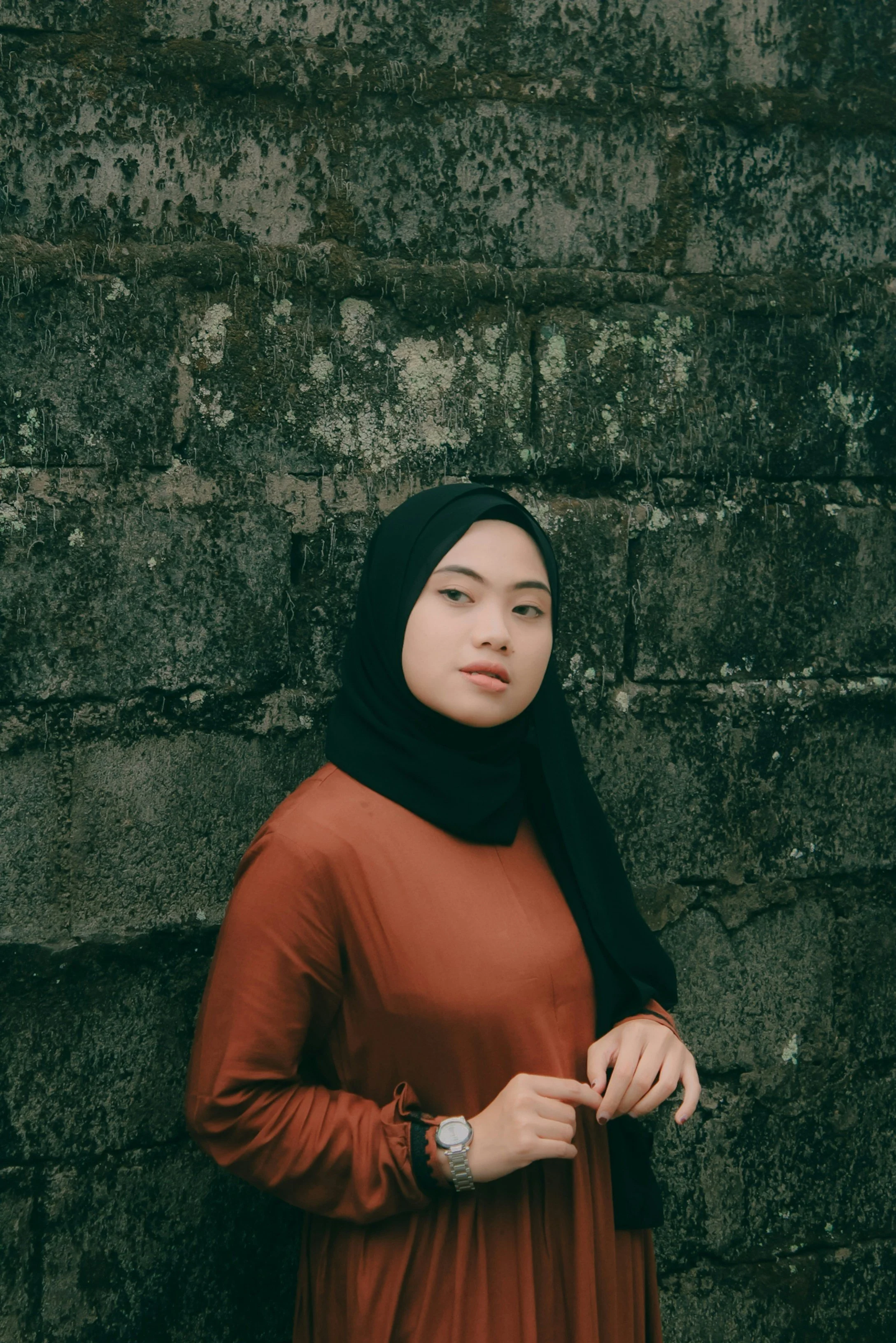 A woman wearing a black headscarf and an orange dress standing against a moss-covered stone wall.