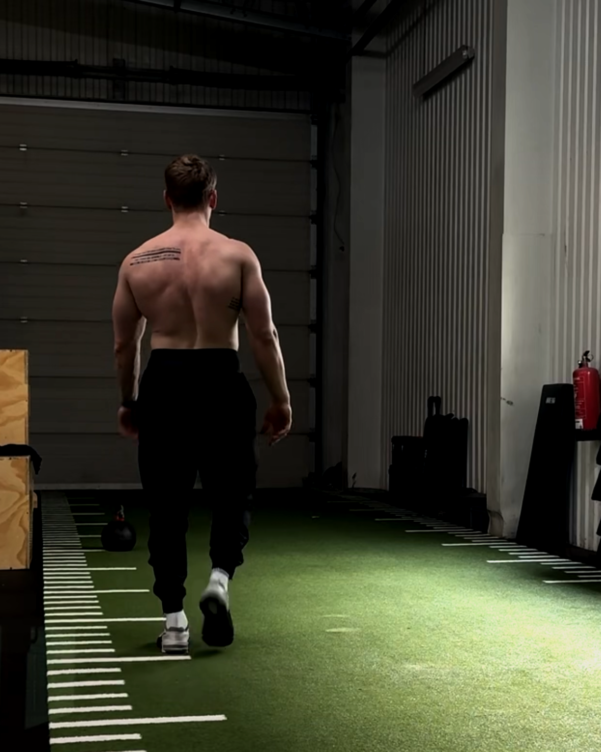Ein Mann trainiert in einer Fitnesshalle, trägt keine Oberbekleidung und geht auf einen markierten Pfad auf grünem Boden.