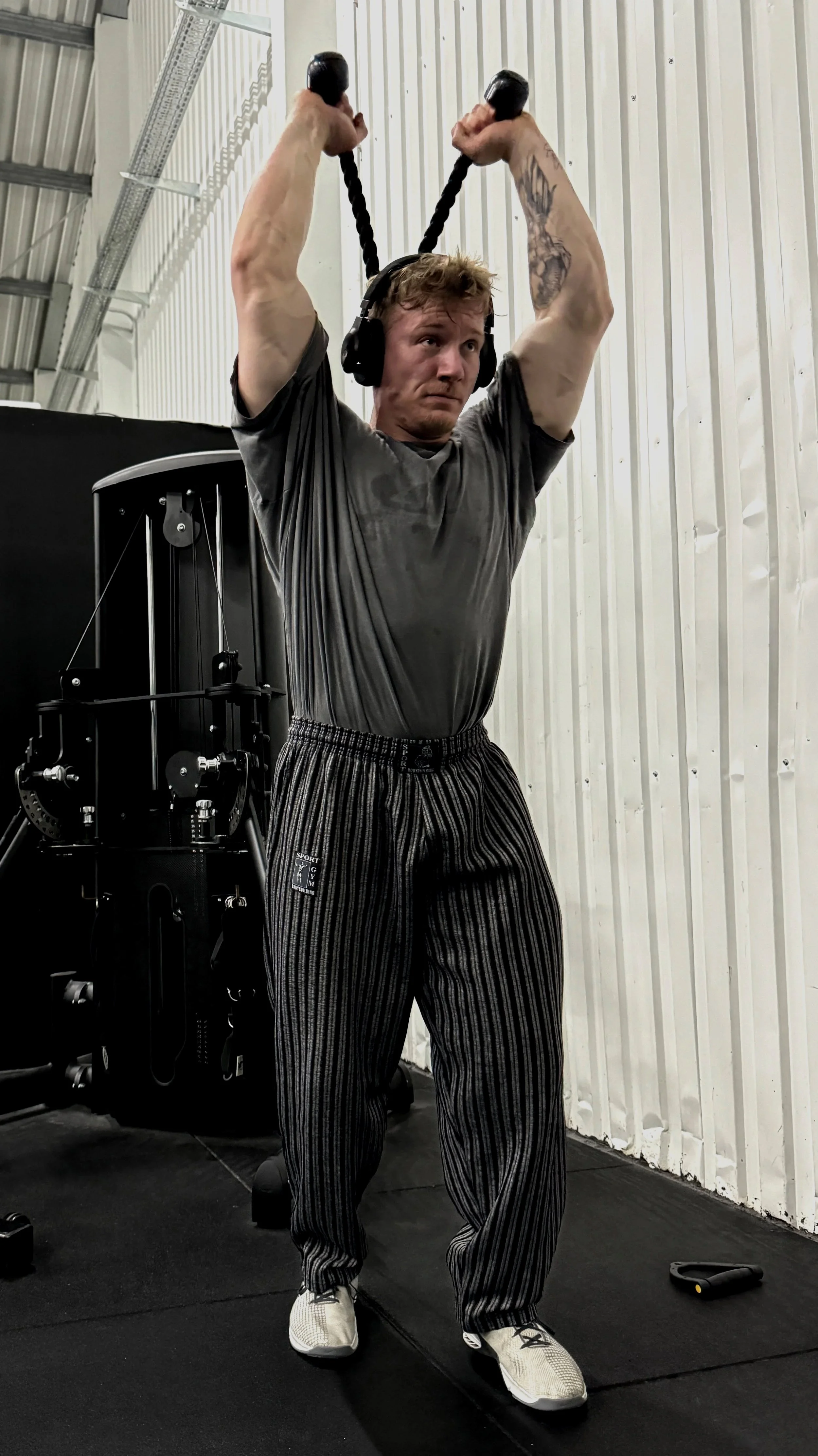 Junger Mann beim Training in einem Fitnessstudio, der eine Schulterübung mit Seilen macht, trägt Kopfhörer, ein graues T-Shirt, schwarze und weiße Turnschuhe und hat Tattoos am Arm.