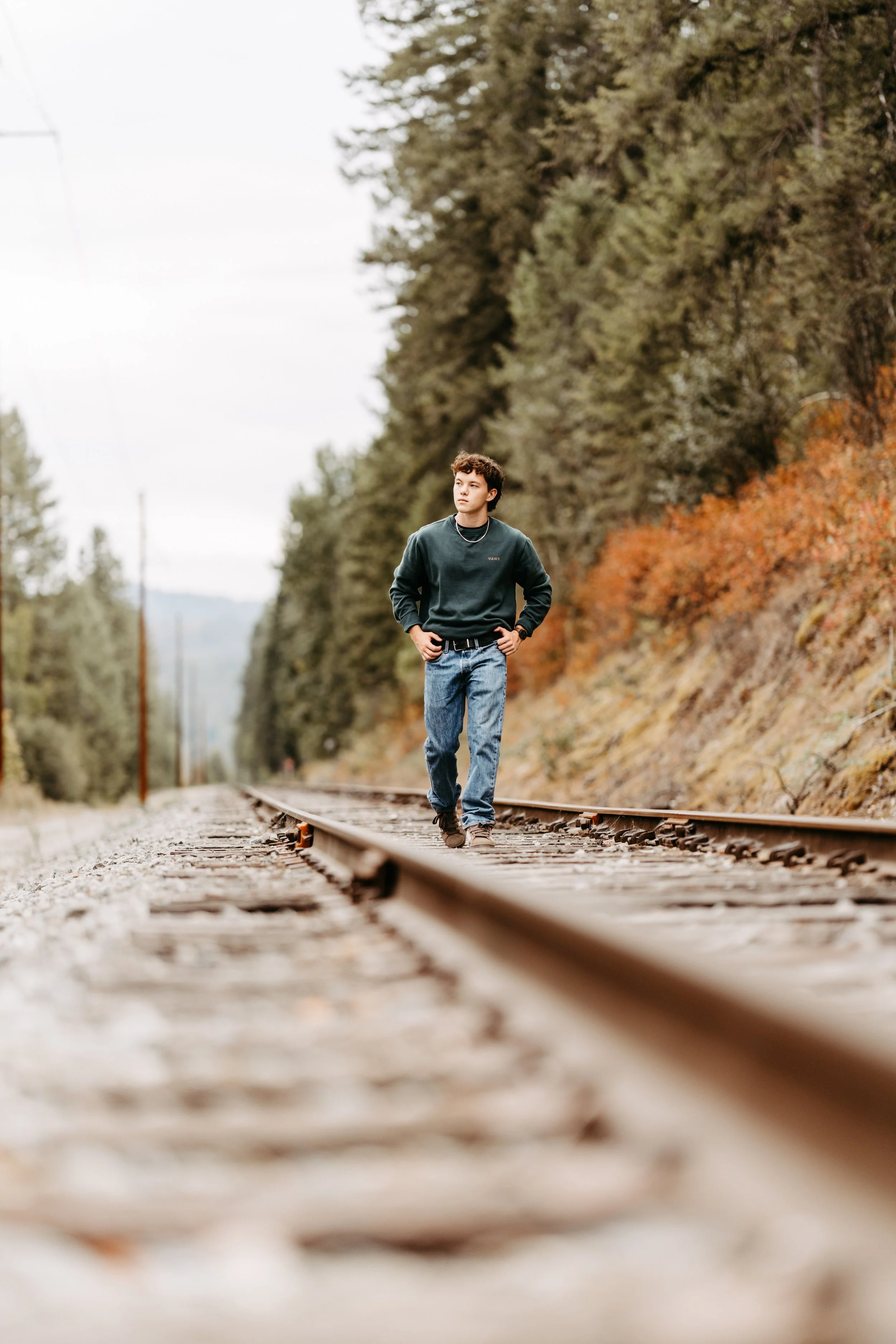 A young man wearing a cowboy hat, glasses, a striped shirt, and jeans, leaning against a wooden fence in an outdoor wooded area, smiling and enjoying himself. Senior photos