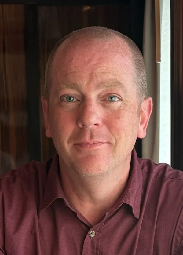 Portrait of a middle-aged man with short, light hair, wearing a maroon button-up shirt, sitting indoors near a window with curtains.