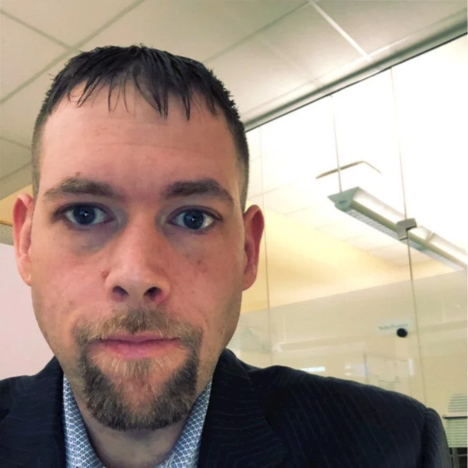 Close-up of a man with short dark hair, blue eyes, a goatee, wearing a dark blazer and light-colored shirt, in an indoor office setting with glass partitions and ceiling lights.