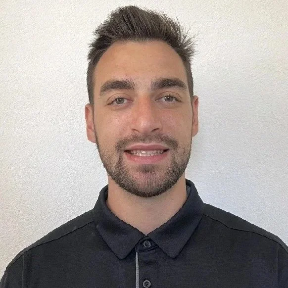 A young man with short, styled brown hair and a beard, smiling, wearing a black polo shirt, standing against a plain white wall.