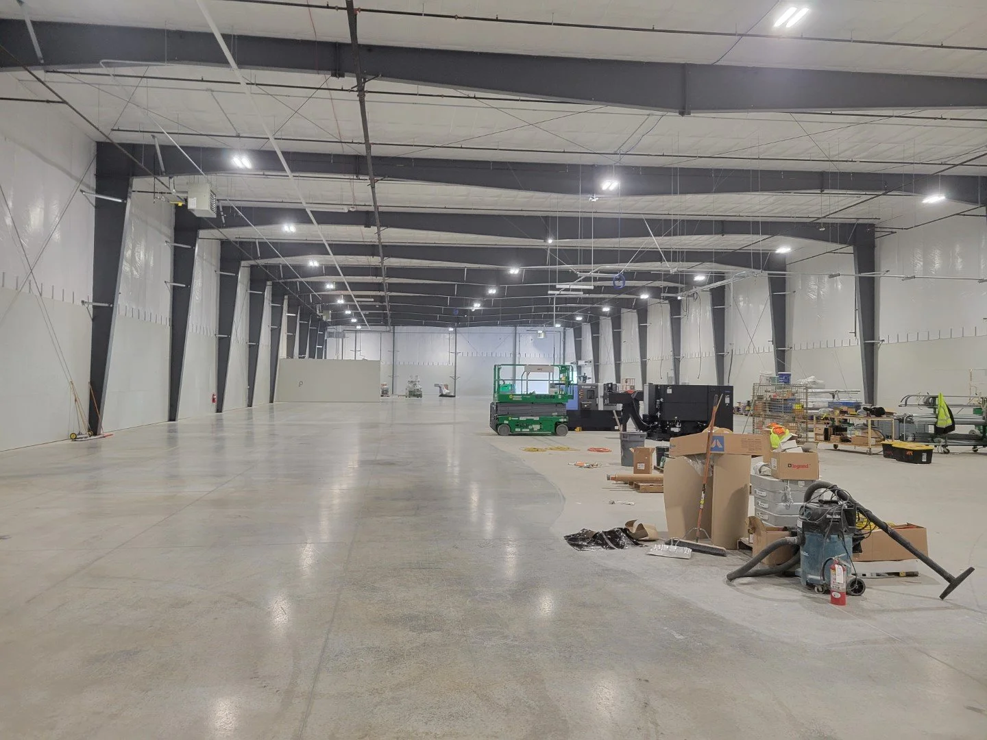 Empty industrial warehouse with various construction and cleaning equipment, tools, and materials scattered on the floor, with a high ceiling and large open space.