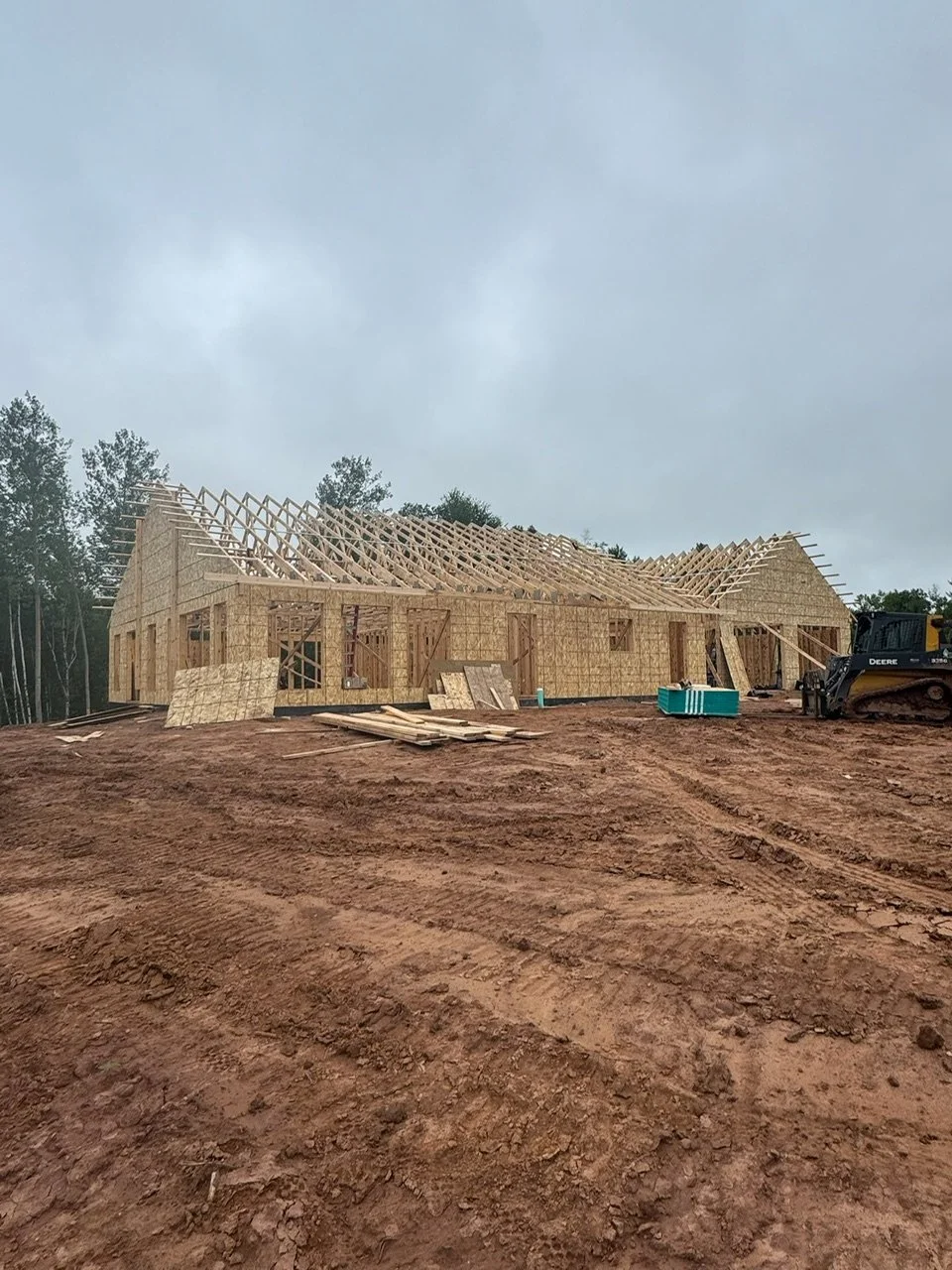 Construction site with a wooden house frame in progress on muddy ground, overcast sky, and construction equipment nearby.
