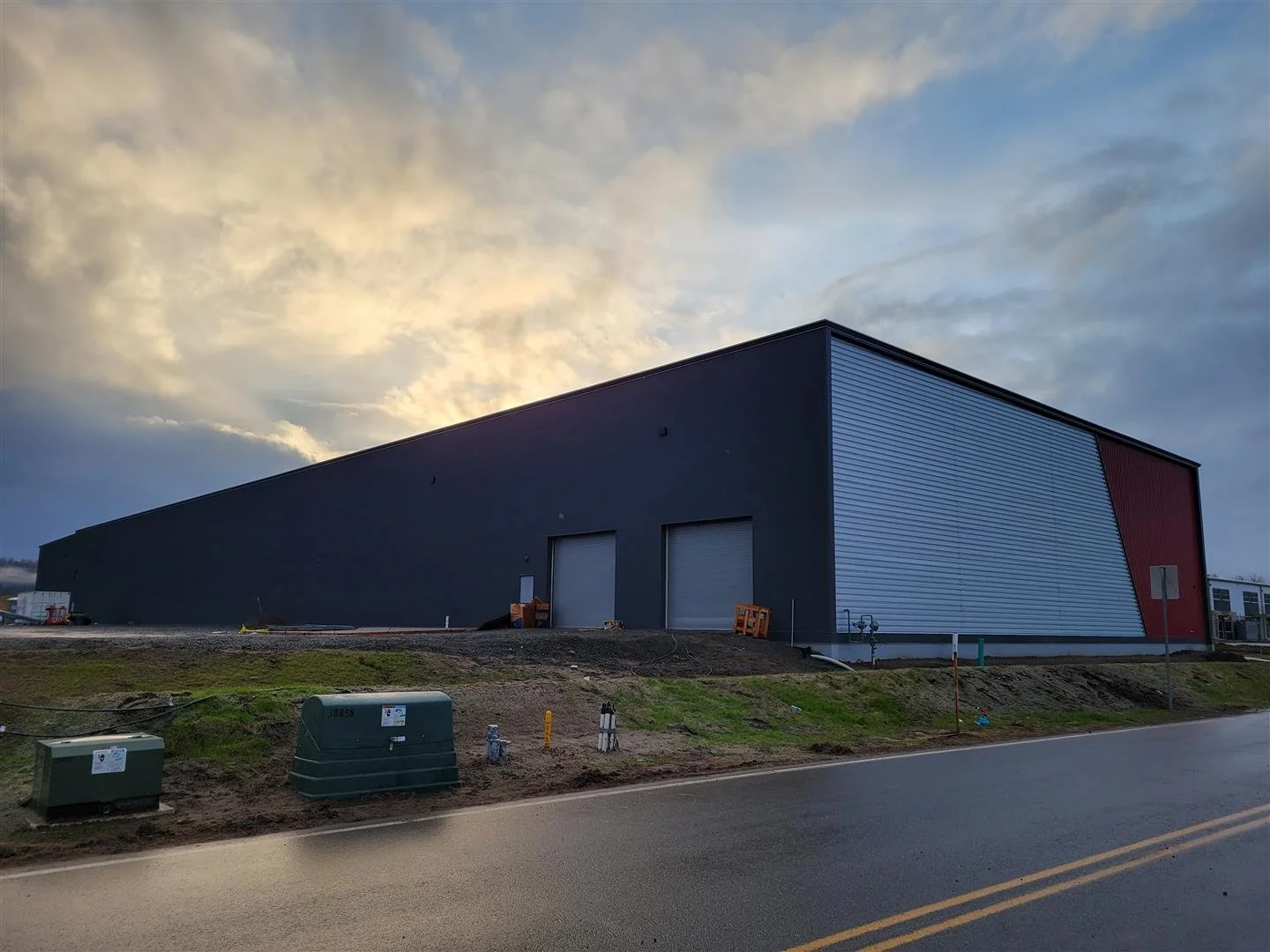 A large modern industrial building with a dark exterior, featuring a distinctive angled metal facade on one side, and two large garage doors. The building is situated along a paved road with some utility boxes and construction materials nearby, and the sky above is partly cloudy during sunset or sunrise.