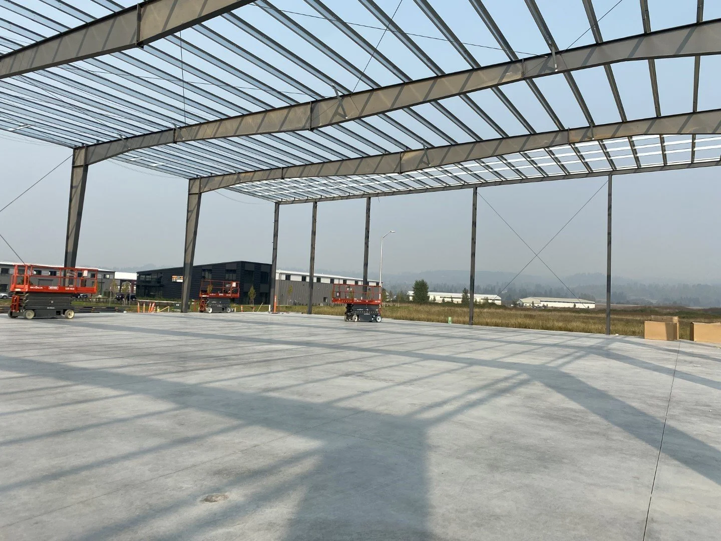 Construction site with a steel framework for a roof structure and scissor lifts, with an open concrete floor and surrounding construction materials.
