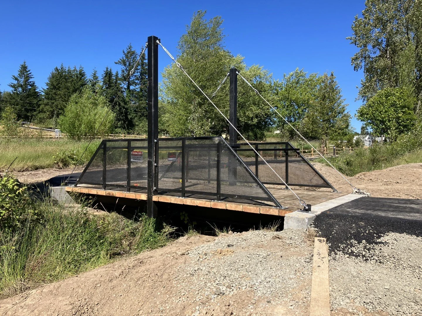 Small bridge with black metal railing and cables, constructed over a dirt and gravel path in a green outdoor area on a clear, sunny day.