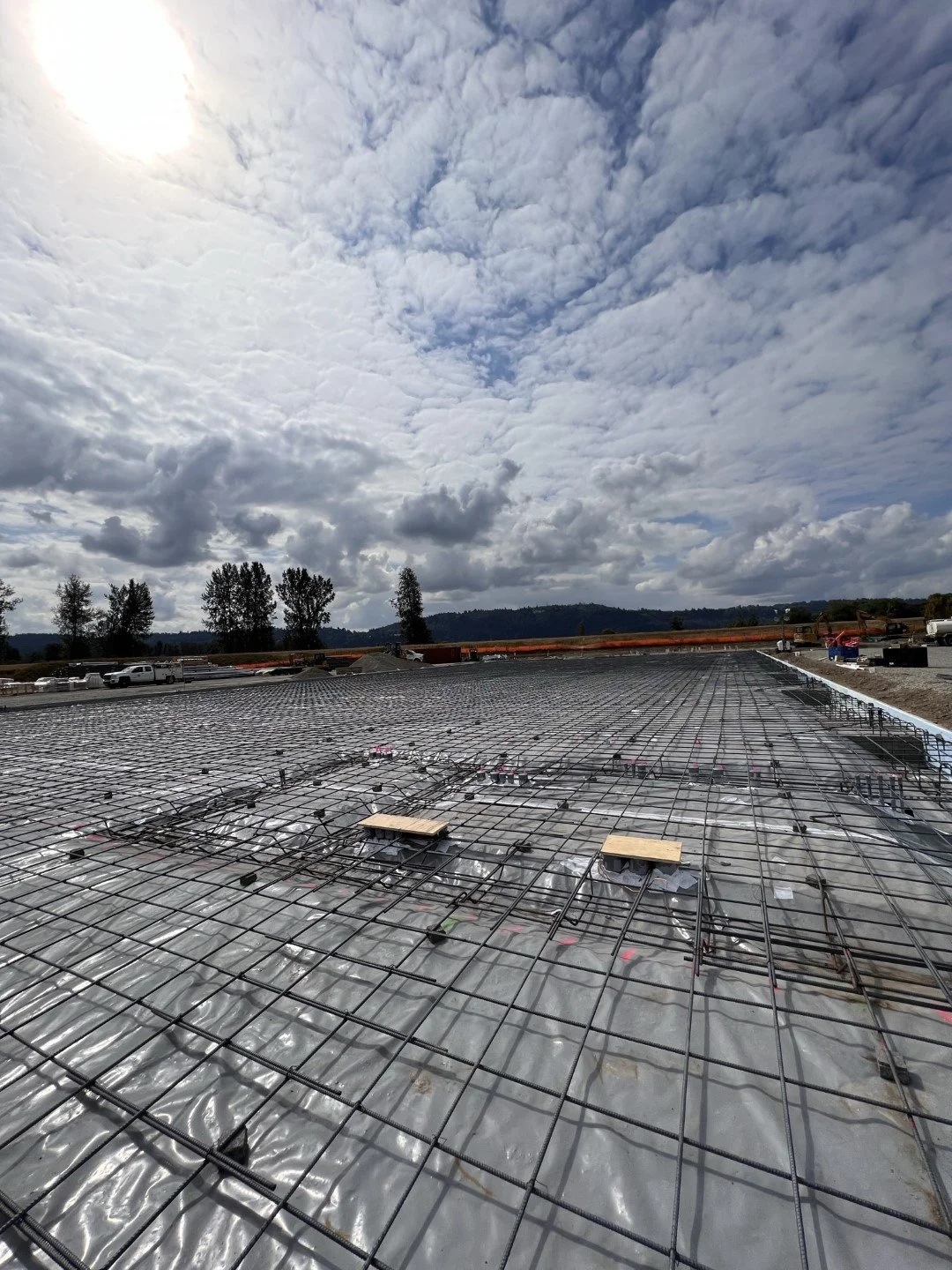 Construction site with a reinforced steel mesh foundation, over a plastic vapor barrier, under partly cloudy sky, with trees and vehicles in the background.