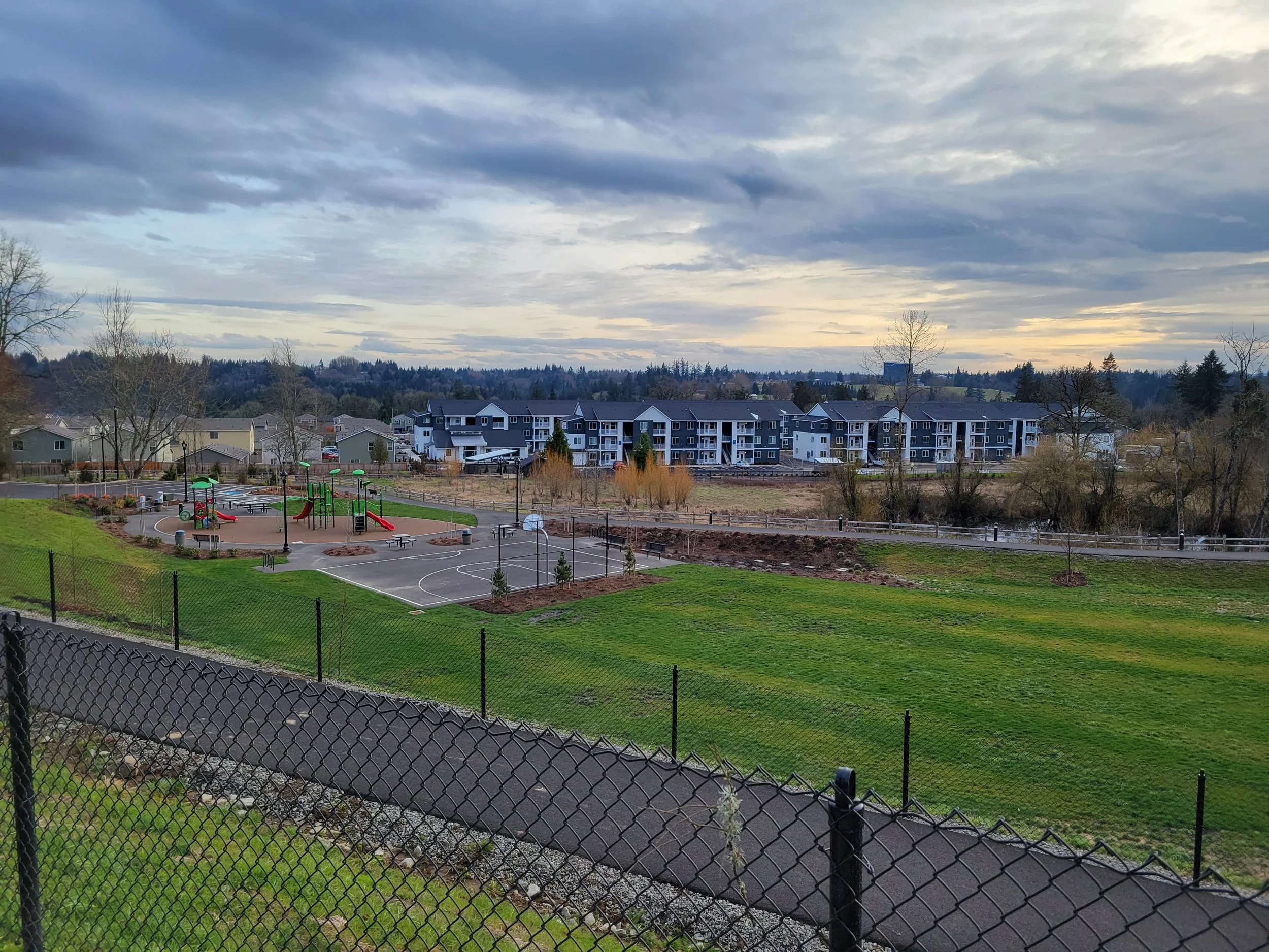 Empty playground and basketball court with apartment buildings and trees in the background on a cloudy day.