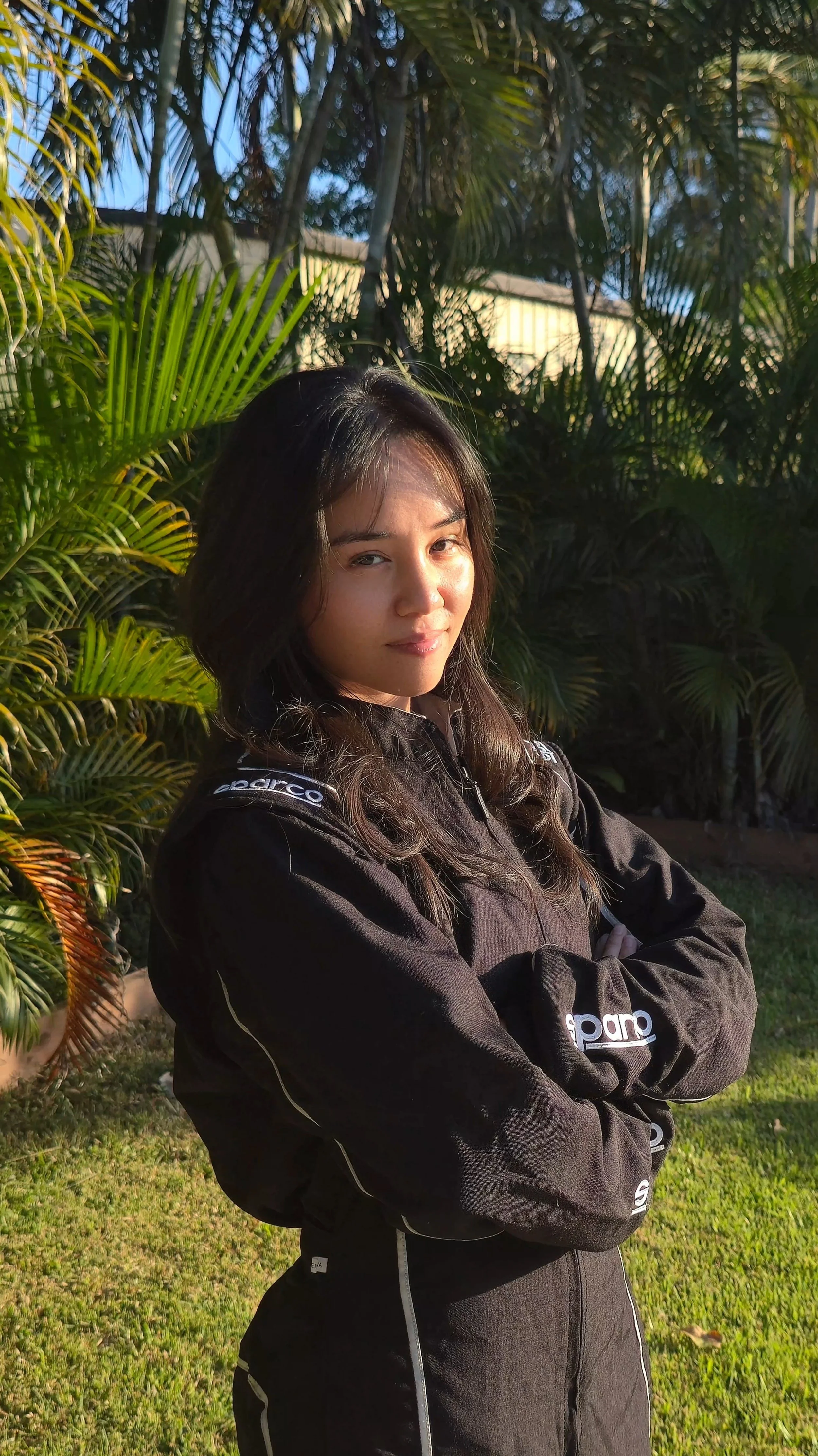X Le standing outdoors in front of green foliage, crossing her arms and looking at the camera with a slight smile, wearing a black racing suit.