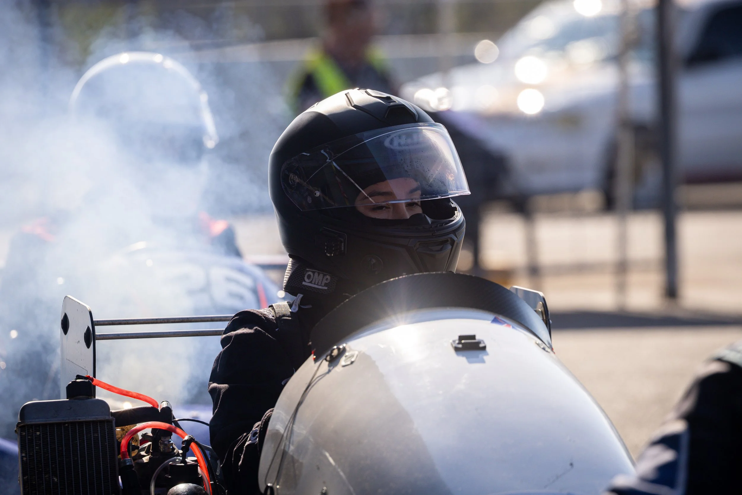 X Le sporting her original unpainted black helmet and racing suit sits in their Rotax Max Superkart, waiting in marshalling before a race start amidst a smoky setting.