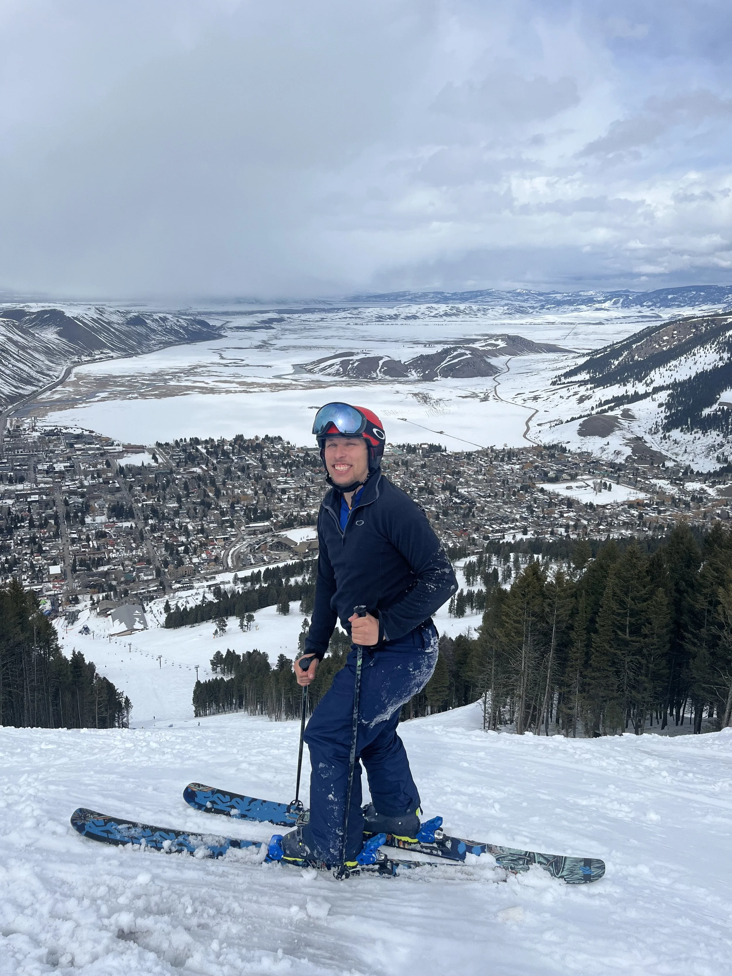 A man wearing a helmet and ski gear stands on a snowy slope with a town, a lake, and snow-covered mountains in the background.