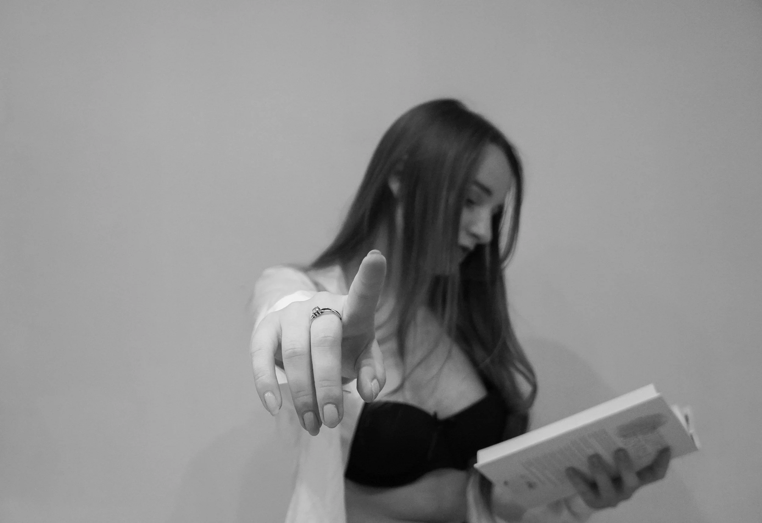 A young woman with long hair is pointing towards the camera with her finger while holding a book in her other hand. She is wearing a black top and a ring and is looking down at the book.