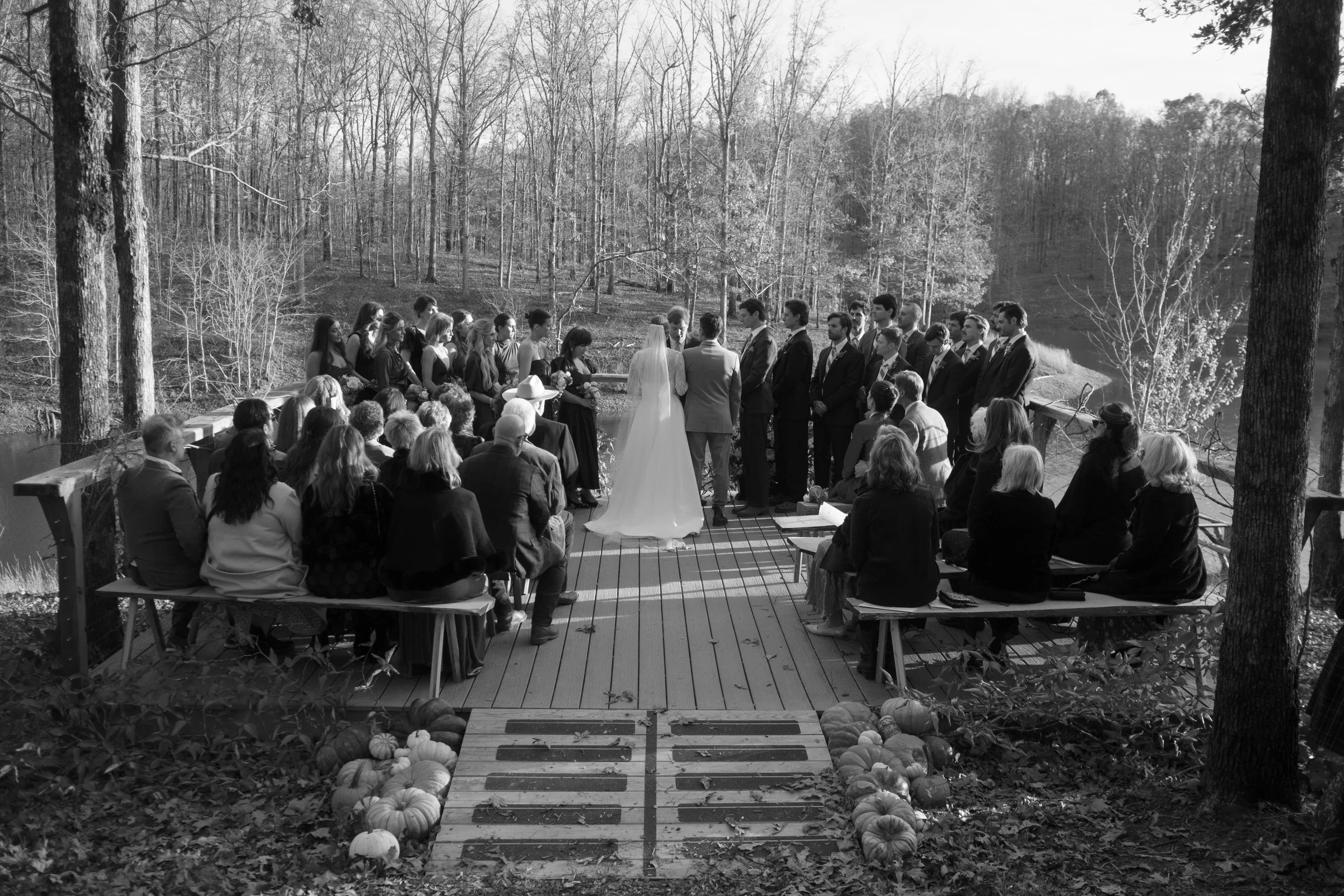 A black and white photo of a wedding ceremony taking place outdoors on a wooden deck surrounded by trees, with guests seated on benches, and the bride and groom standing at the altar.