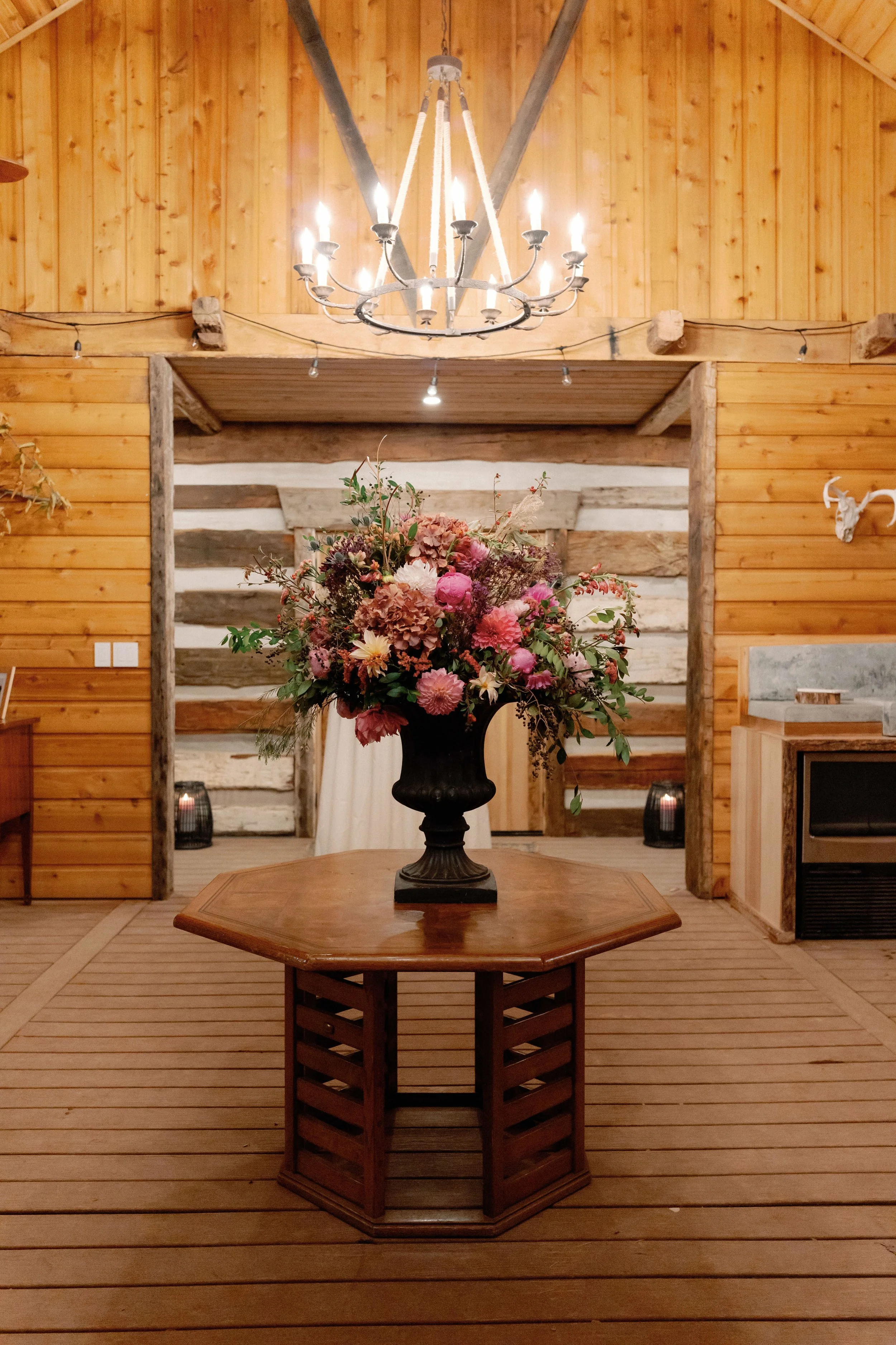 A rustic wooden interior with a central floral arrangement on a wooden table, a chandelier hanging from the ceiling, and warm lighting.