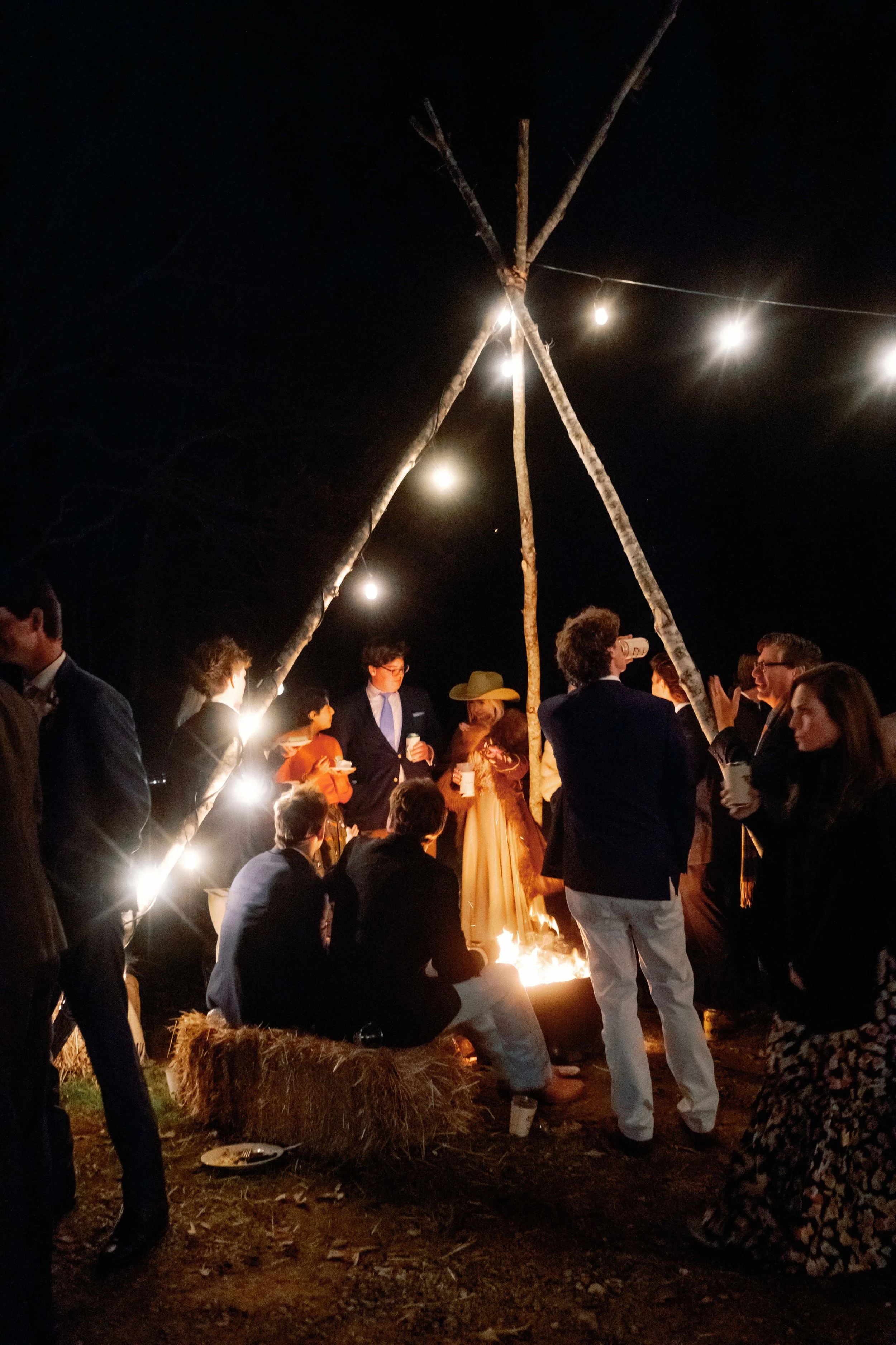 Nighttime outdoor gathering around a fire pit with string lights overhead, people dressed in formal and casual attire standing and sitting, some holding drinks.