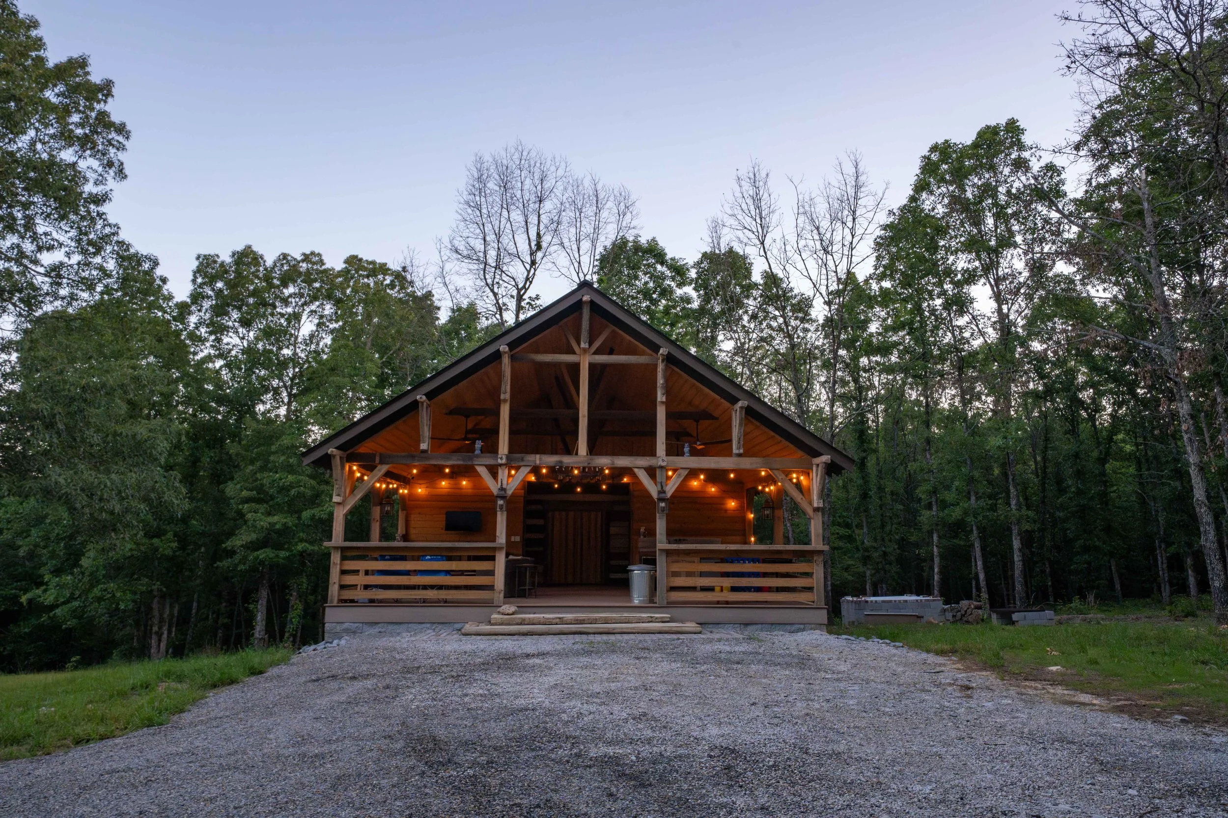 A wooden cabin with string lights on the porch, surrounded by trees at dusk.