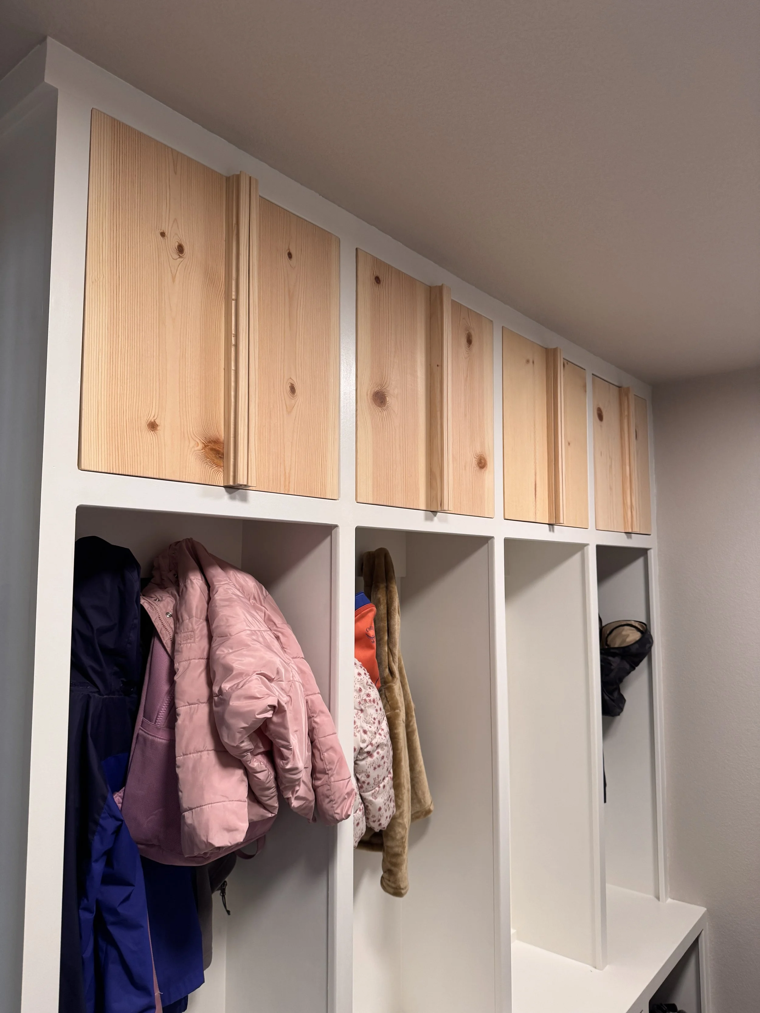 Custom handmade pine cabinet doors installed on white mudroom cubby system with vertical wood handles and open coat storage