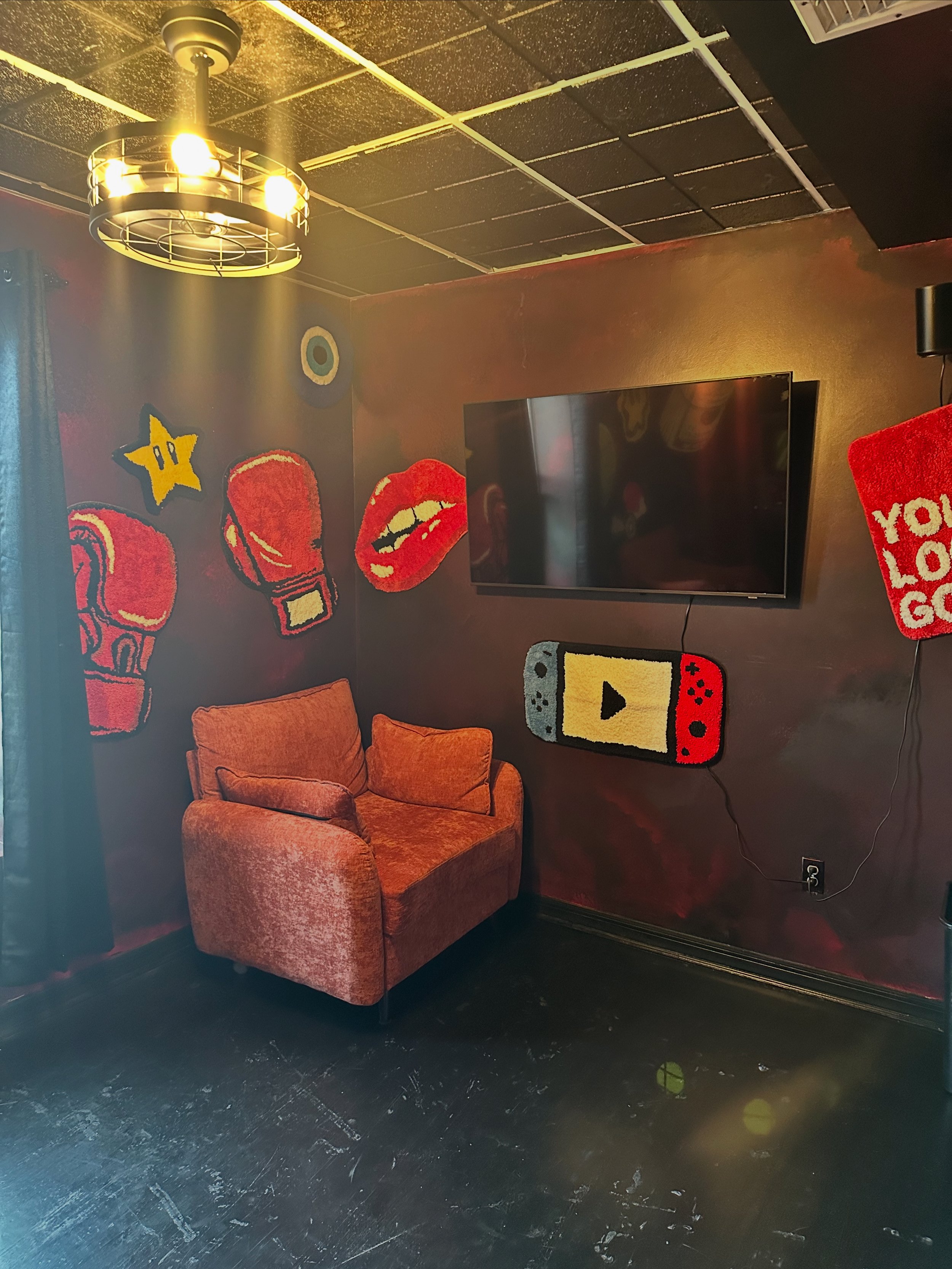 Cozy room with a large television mounted on a red wall decorated with gaming and pop culture pixel art, including a star, boxing gloves, a pair of lips, a game controller-shaped wall hanging, and a red cushion that says 'you look good'. An orange ar