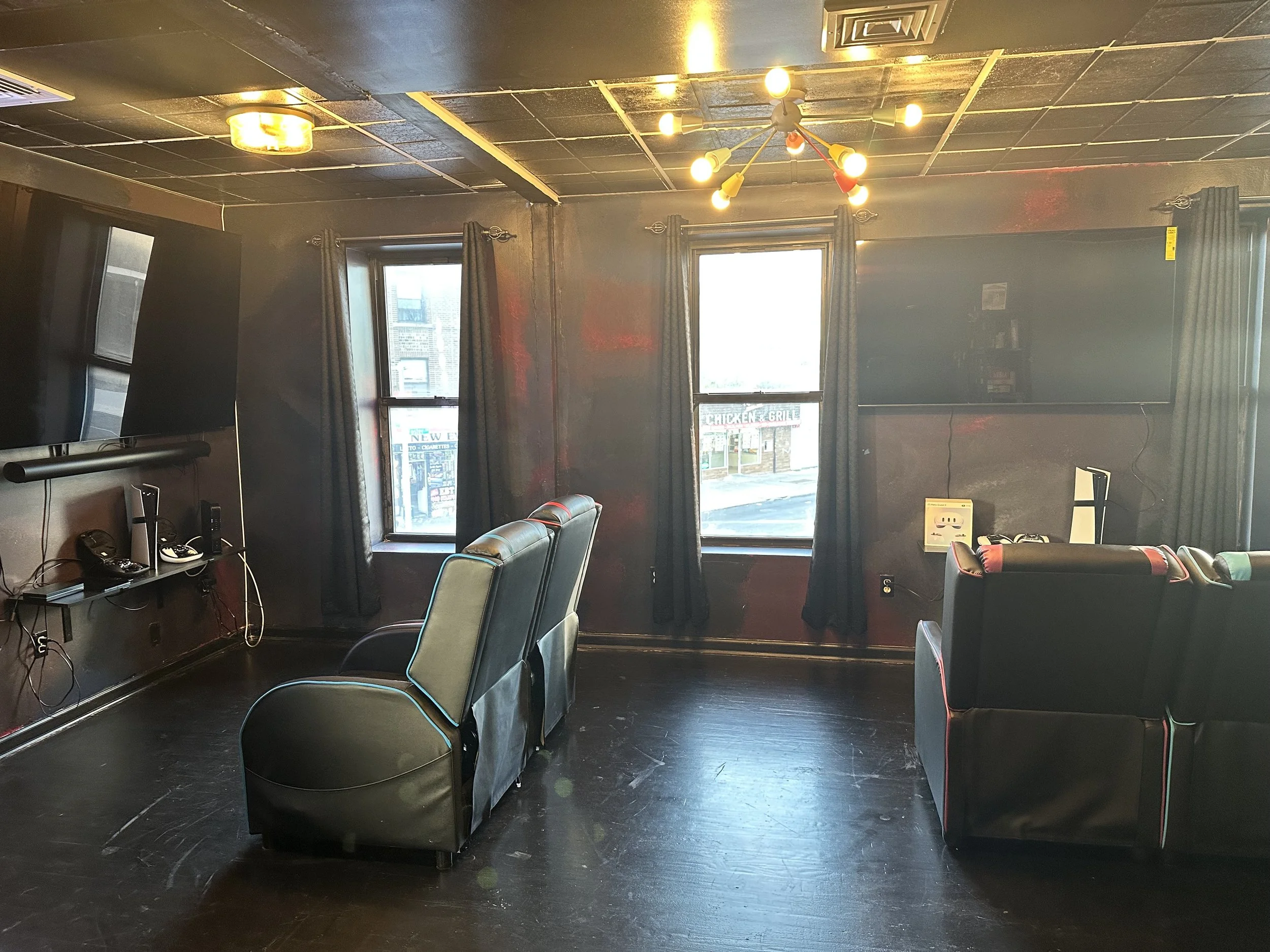 A cozy gaming chairs with two black armchairs, black curtains on windows, a black wall-mounted TV, and a modern ceiling light fixture.