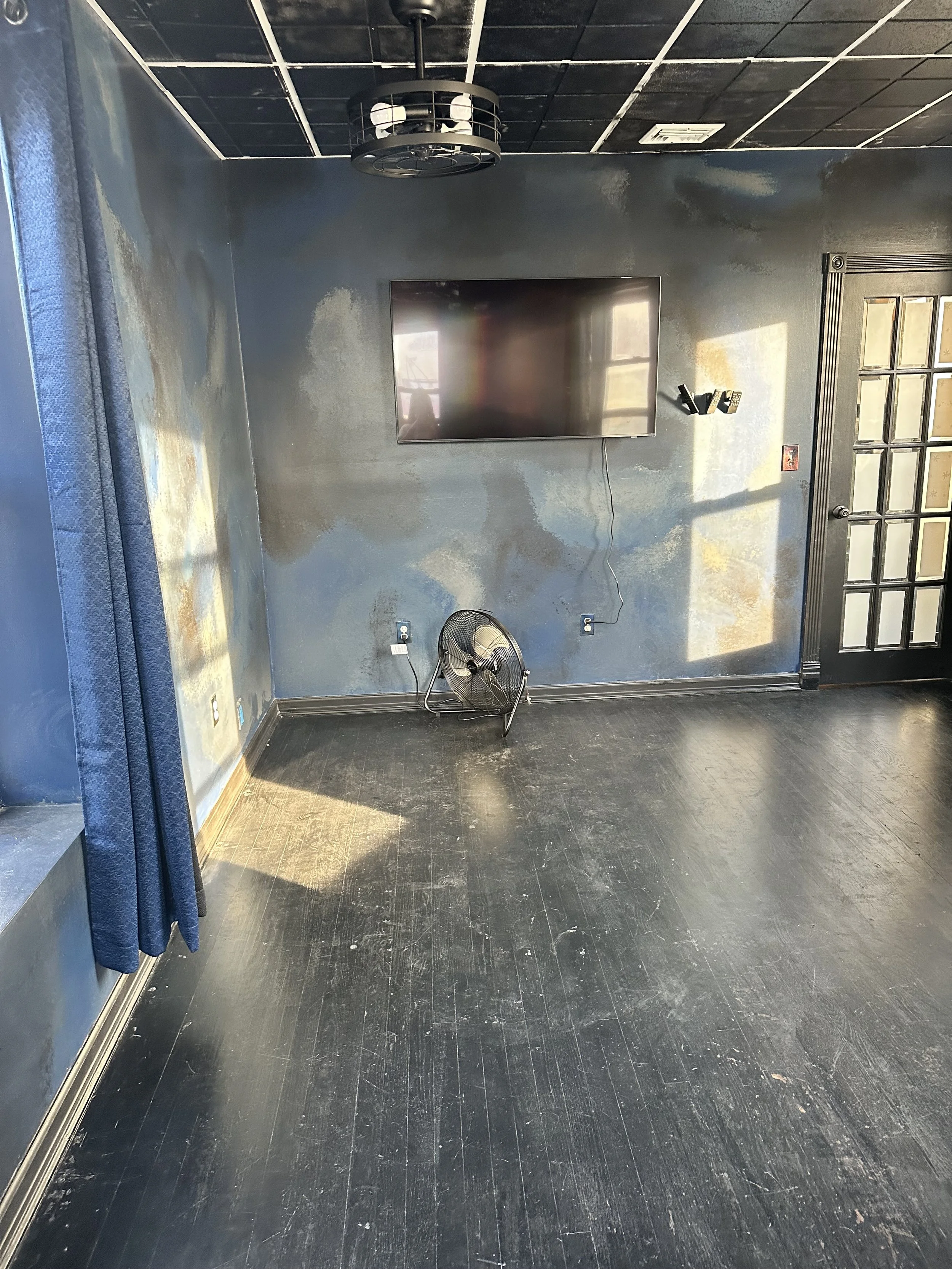 Room with black hardwood floor and dark blue painted walls. Mounted flat-screen TV on wall, with a wire hanging down. Small fan on the floor near the wall. Ceiling is black with grid pattern, ceiling light fixture. Part of a window with blue curtains