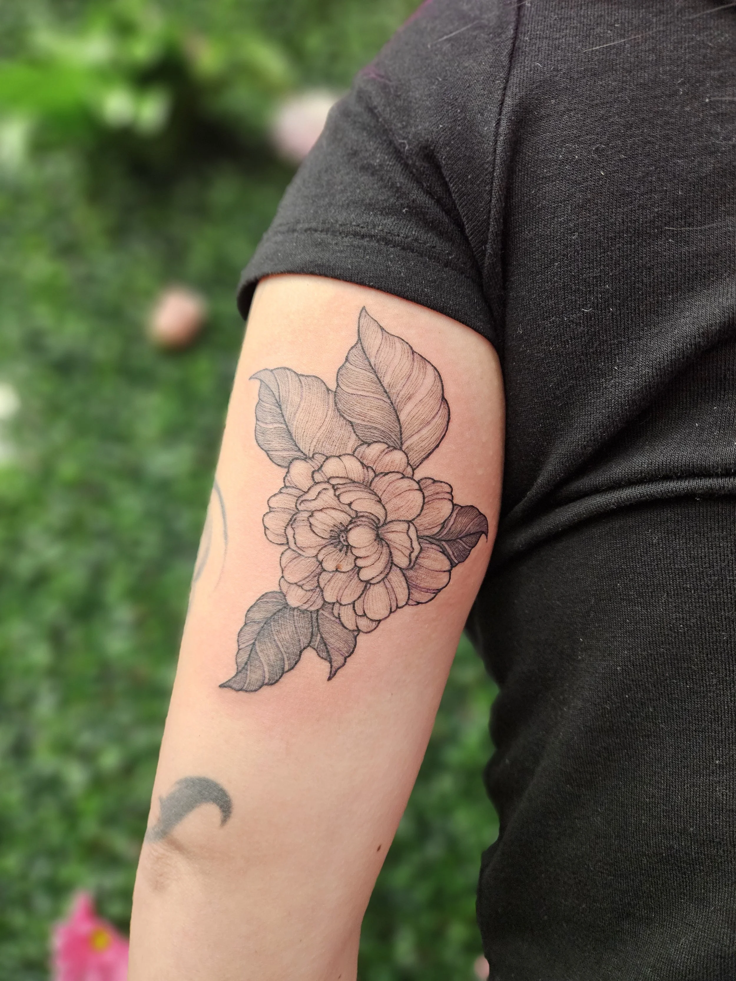 Black and gray floral tattoo on person's upper arm, featuring a large blooming flower with leaves.