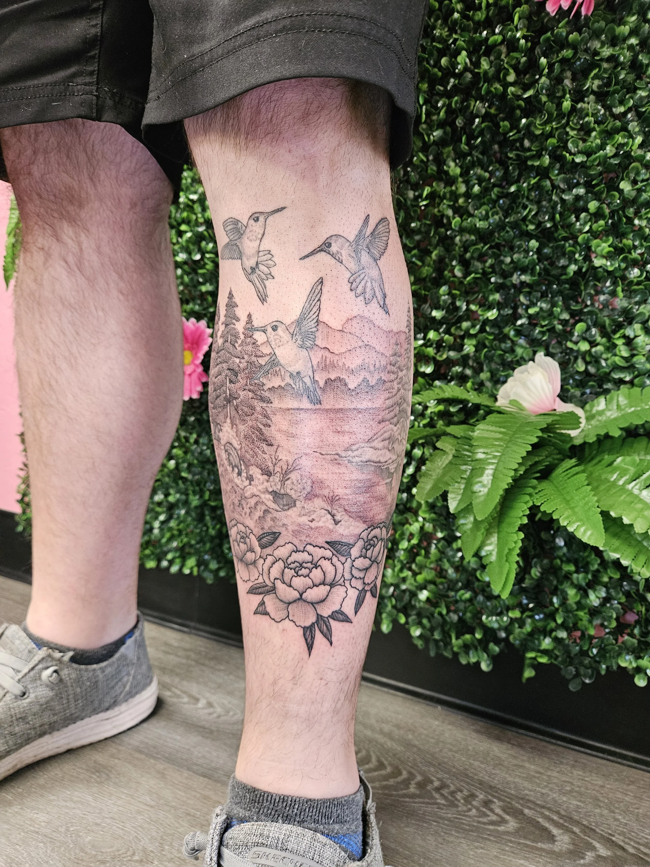 Tattoo of three hummingbirds flying above a landscape with mountains, trees, and flowers on a person's leg.