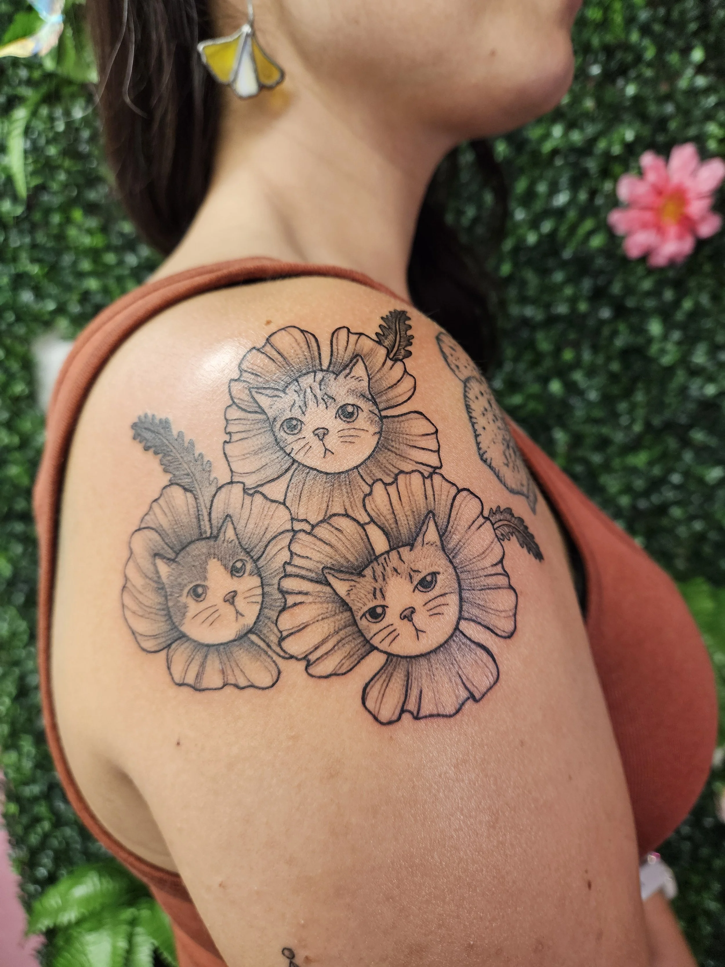 Tattoo of three cat faces with flower petals and leaves on woman's shoulder.