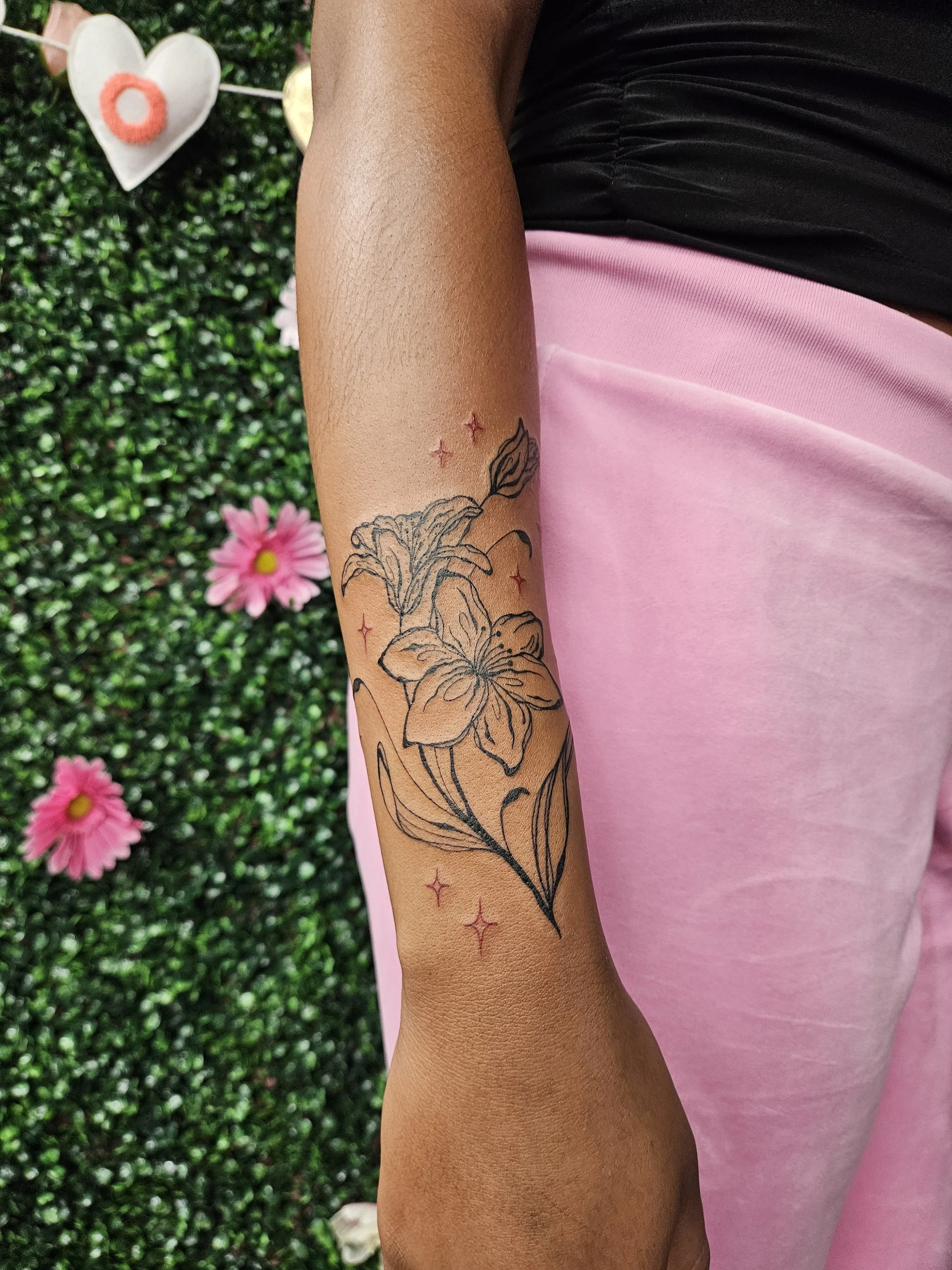 A person's arm with a floral tattoo featuring a lilies, surrounded by small red stars. The person is wearing a pink pants and a black top. In the background, there are pink daisies, a heart-shaped decoration, and a green hedge.