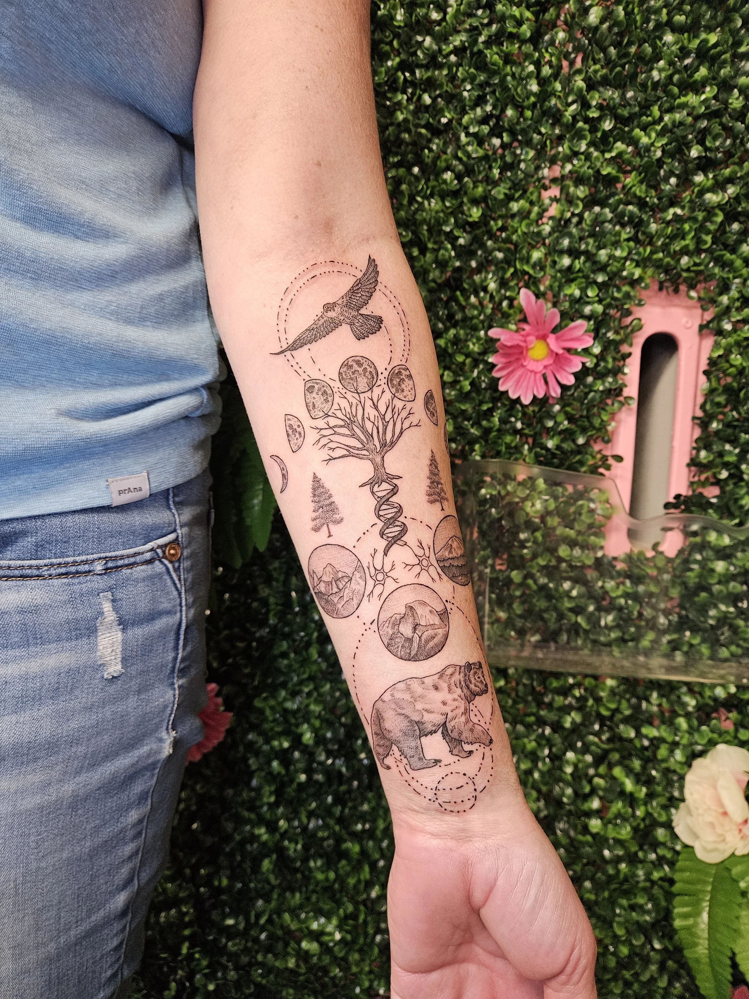 Tattoo on person's forearm featuring a flying bird, a DNA double helix, a bear, mountains, trees, and various geometric shapes and patterns.