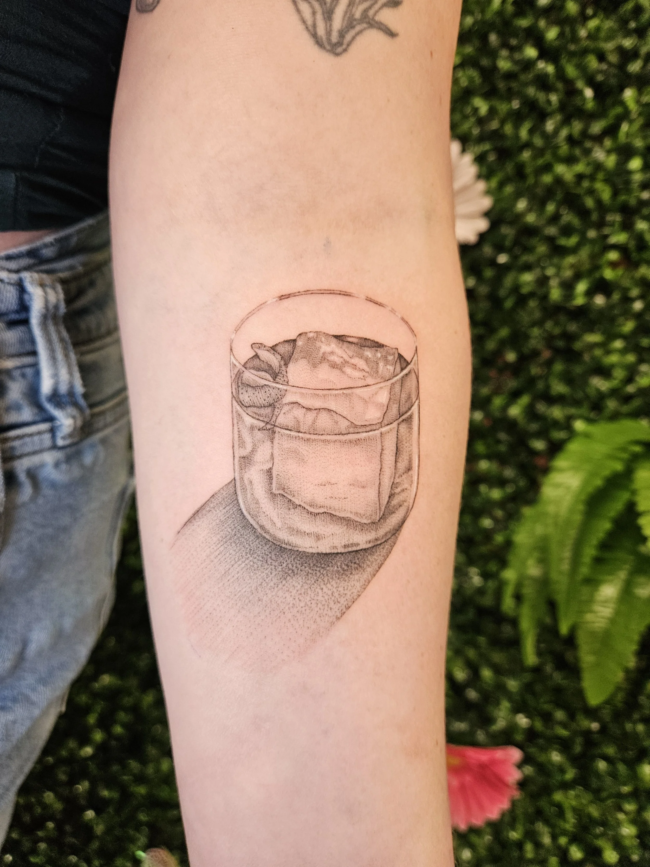 A realistic black and gray tattoo of a glass filled with ice and a beverage, located on a person's forearm.