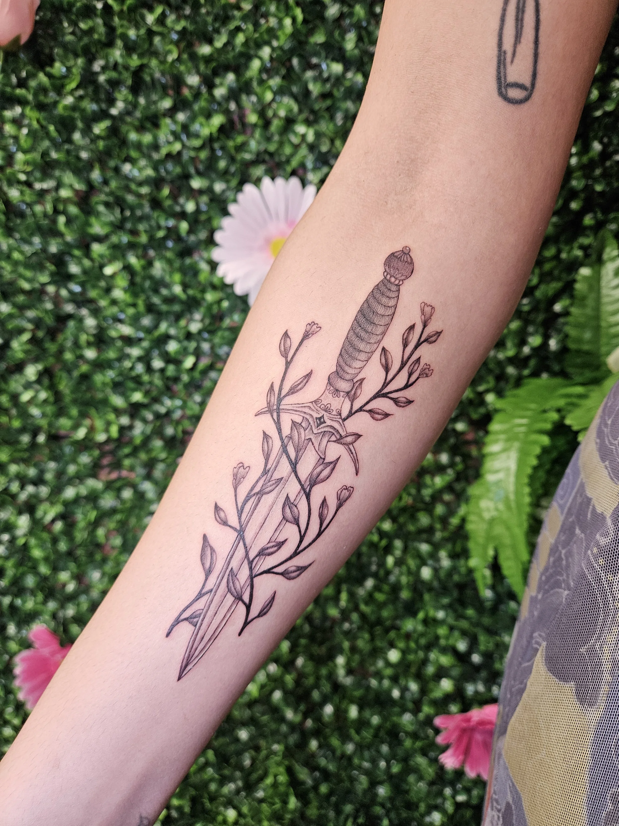 Tattoo of a sword with a scabbard and a decorative handle, surrounded by leaves and small flowers, on a person's forearm.