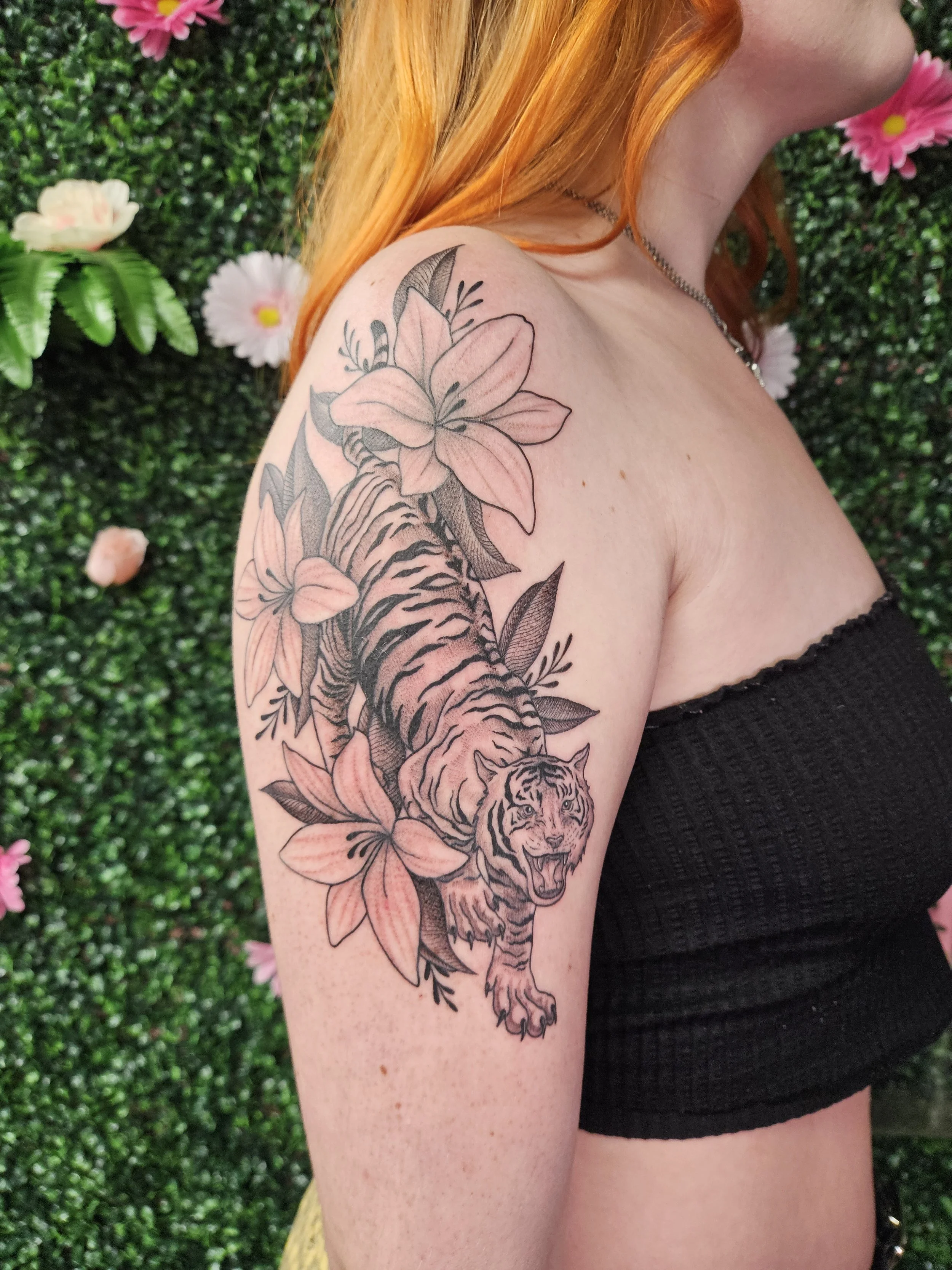 Tattoo of a fierce tiger emerging from pink lilies on woman's upper arm with a green leafy background.