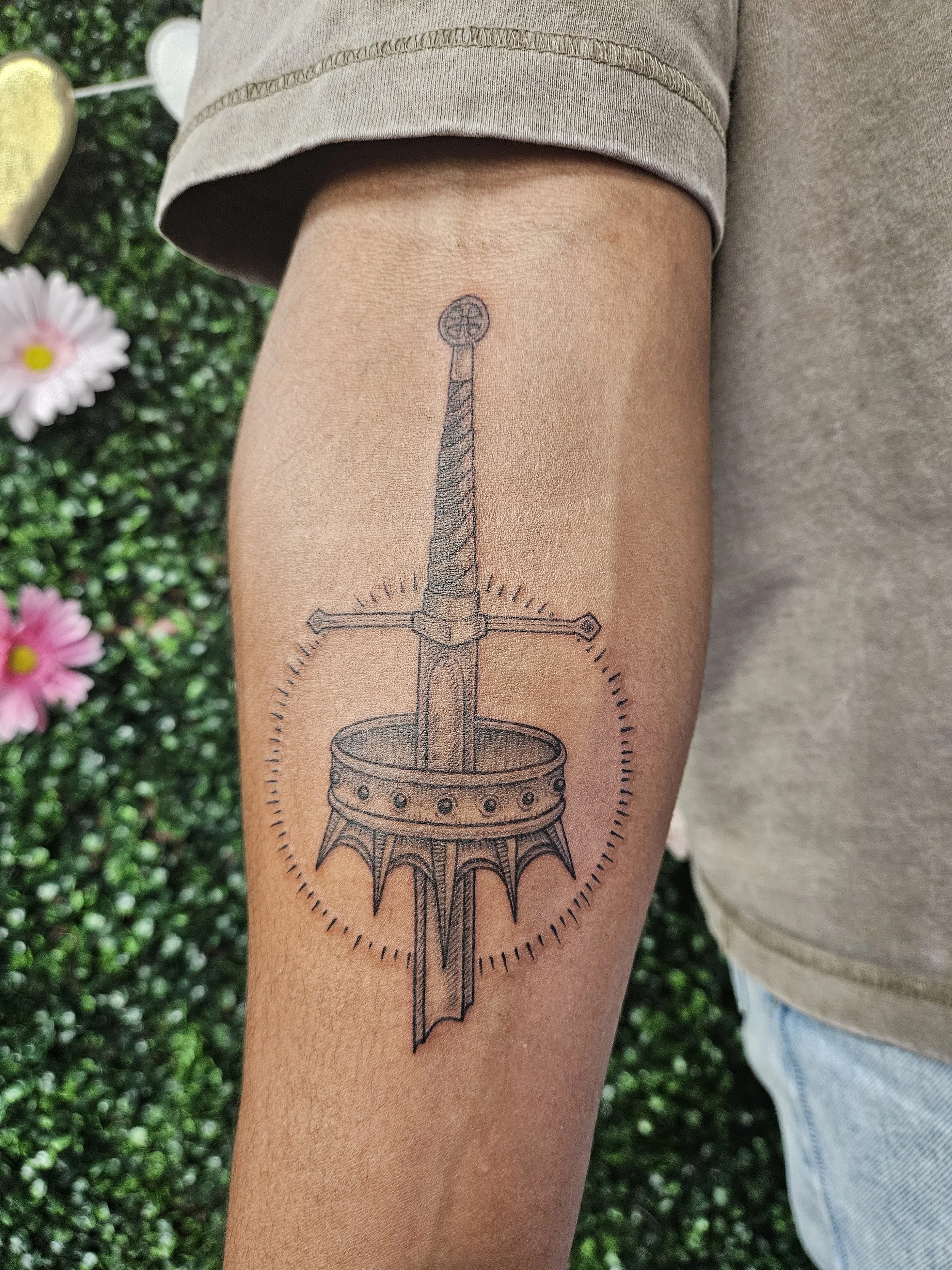 Black and gray tattoo of a sword piercing through a crown on person's upper arm.