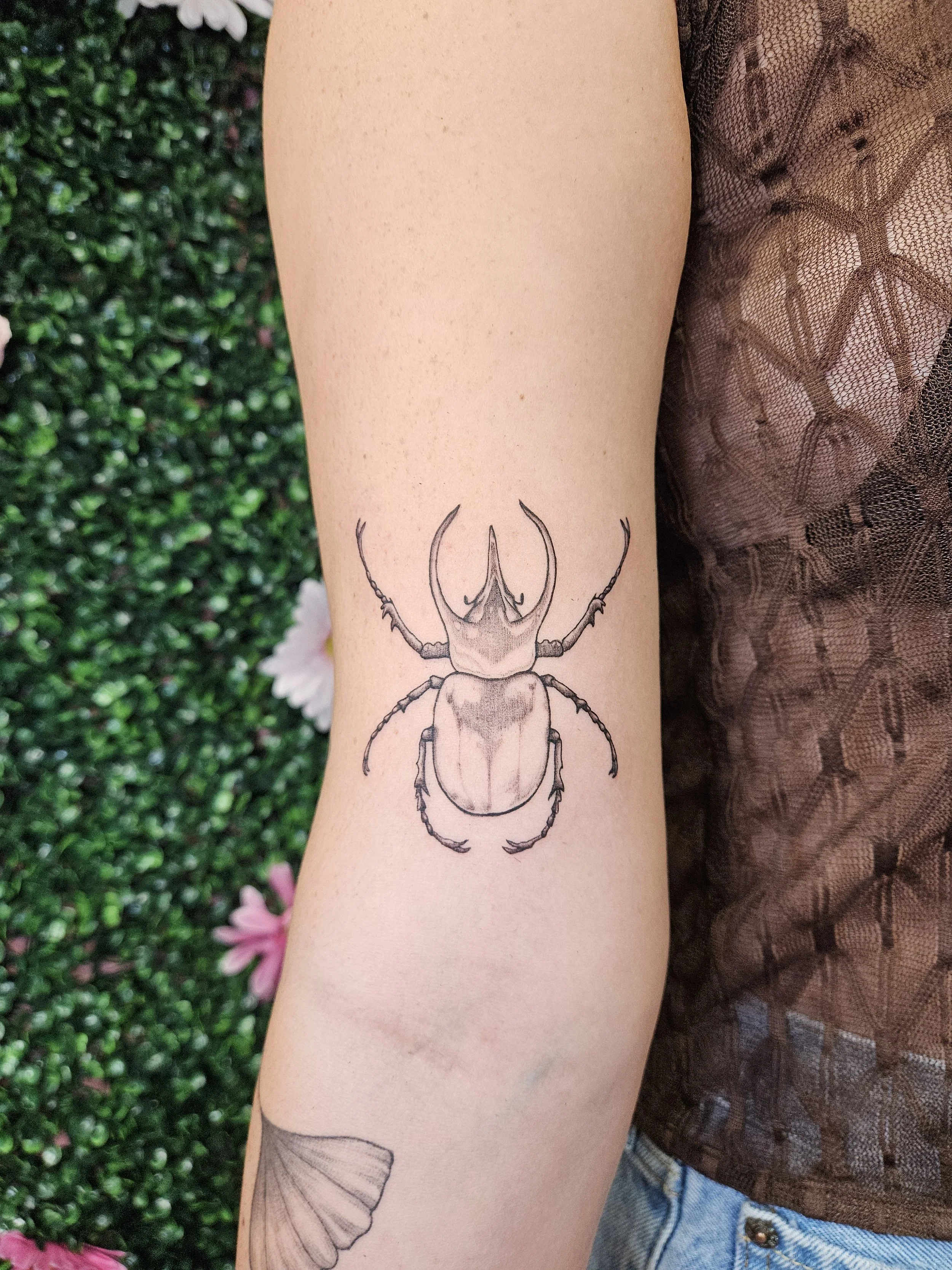 A detailed black and gray tattoo of a beetle on a person's forearm, with a green leafy background and some pink flowers.