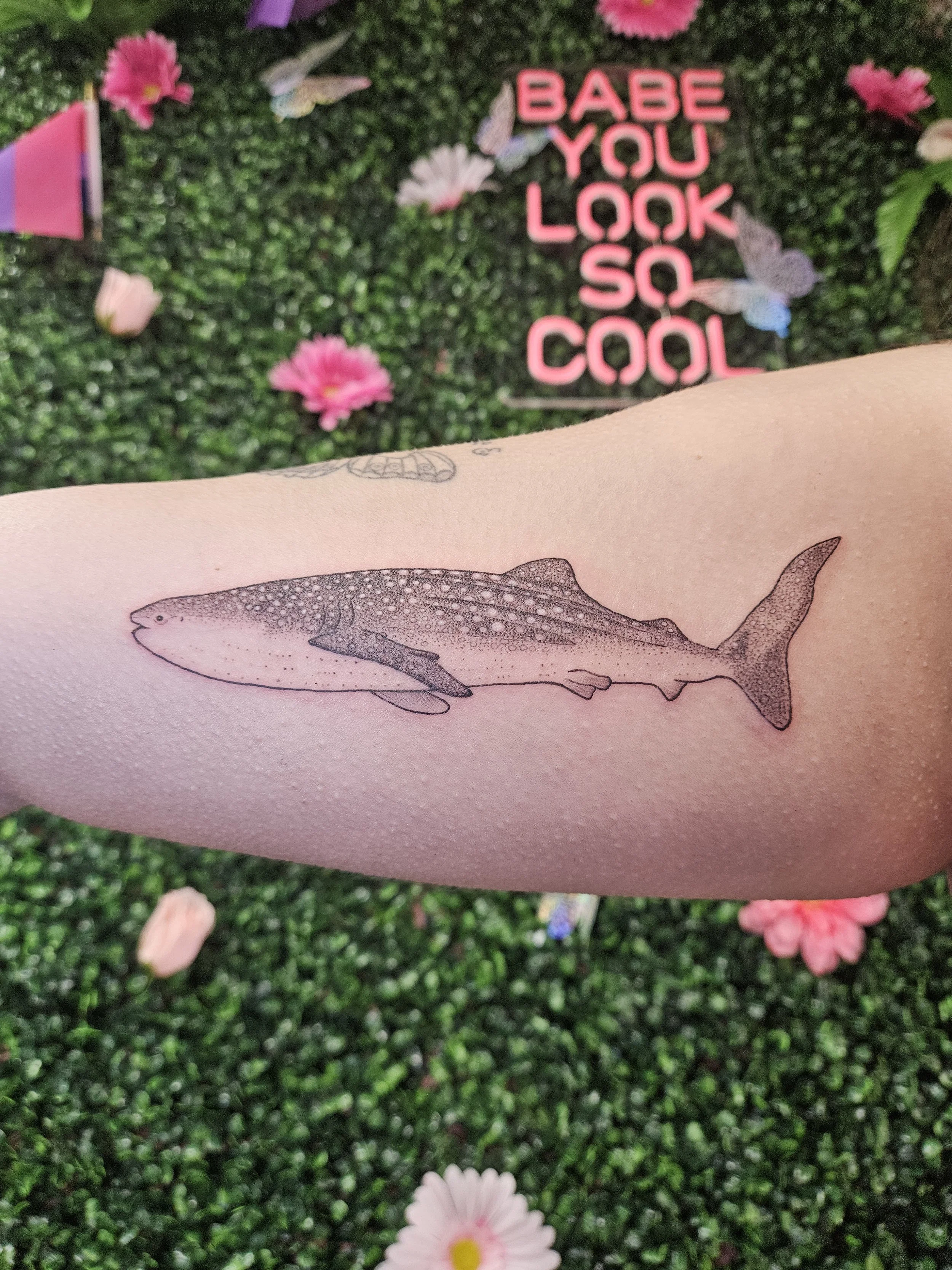 Tattoo of a whale shark on a person's arm, with pink flowers and butterflies in the background. There is a pink sign that says "BABE YOU LOOK SO COOL" among flowers and greenery.