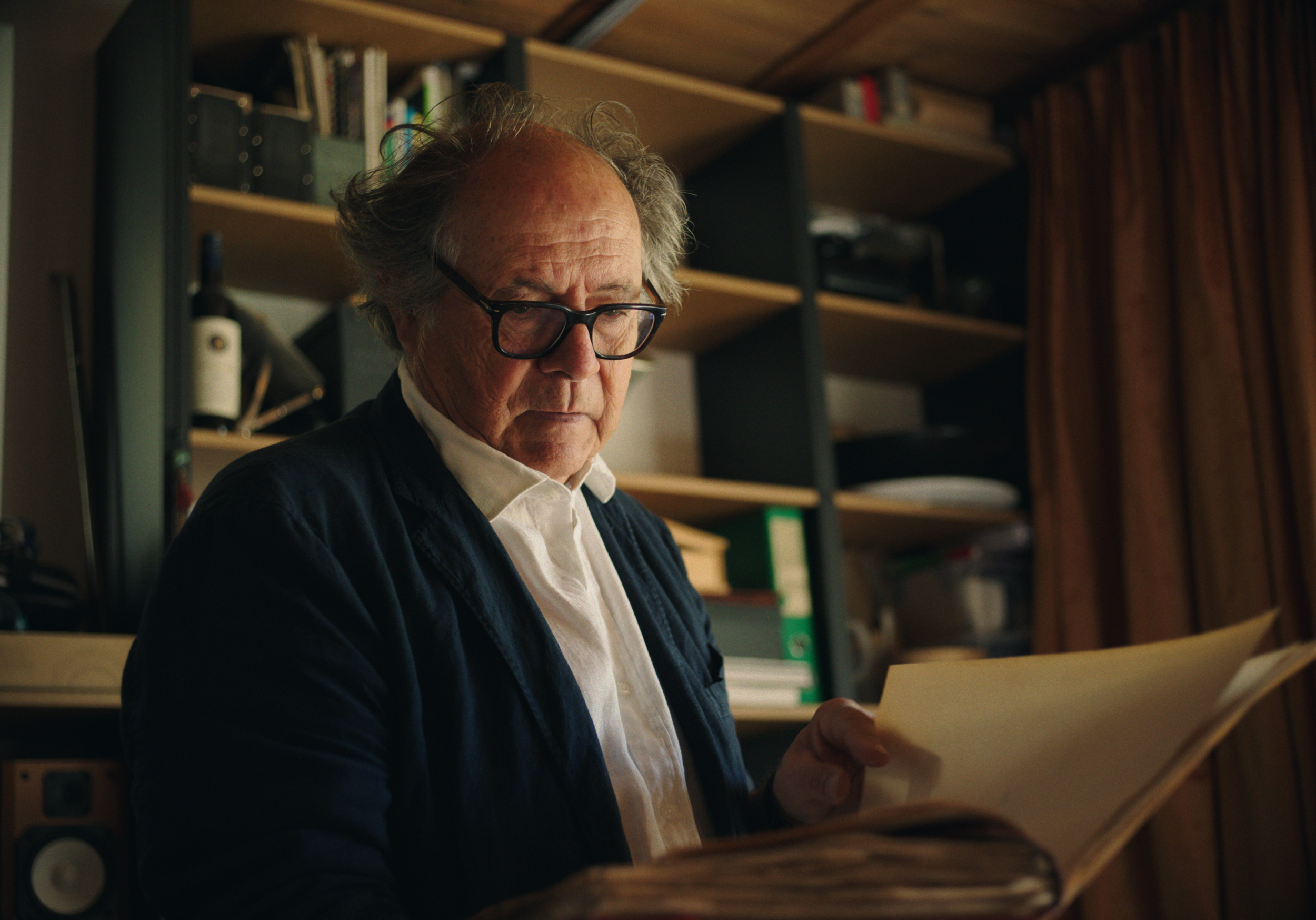 An elderly man with glasses reads a large folder or book in a room with wooden bookshelves and brown curtains.