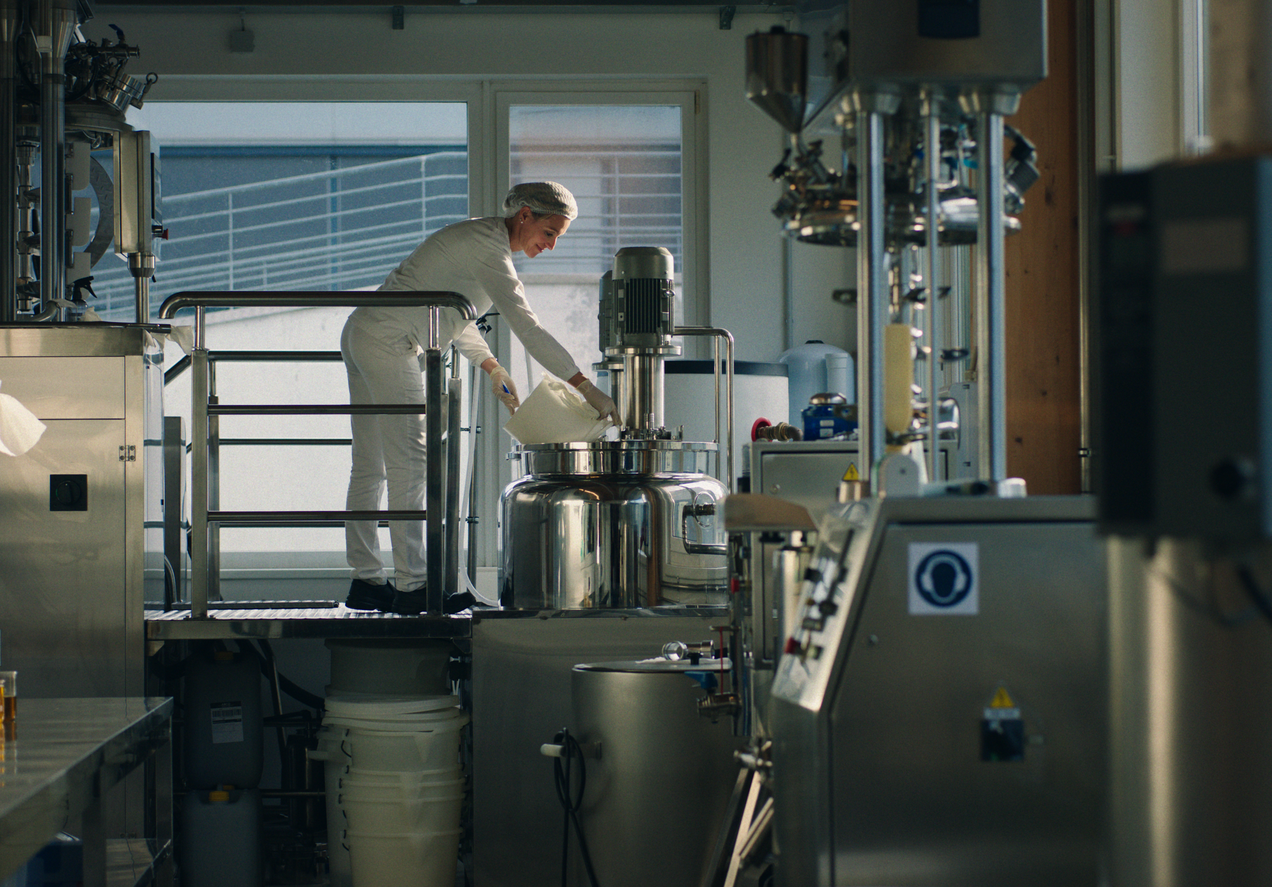 A person in white protective clothing working in a clean industrial laboratory with stainless steel equipment, pouring a liquid into a large stainless steel container.