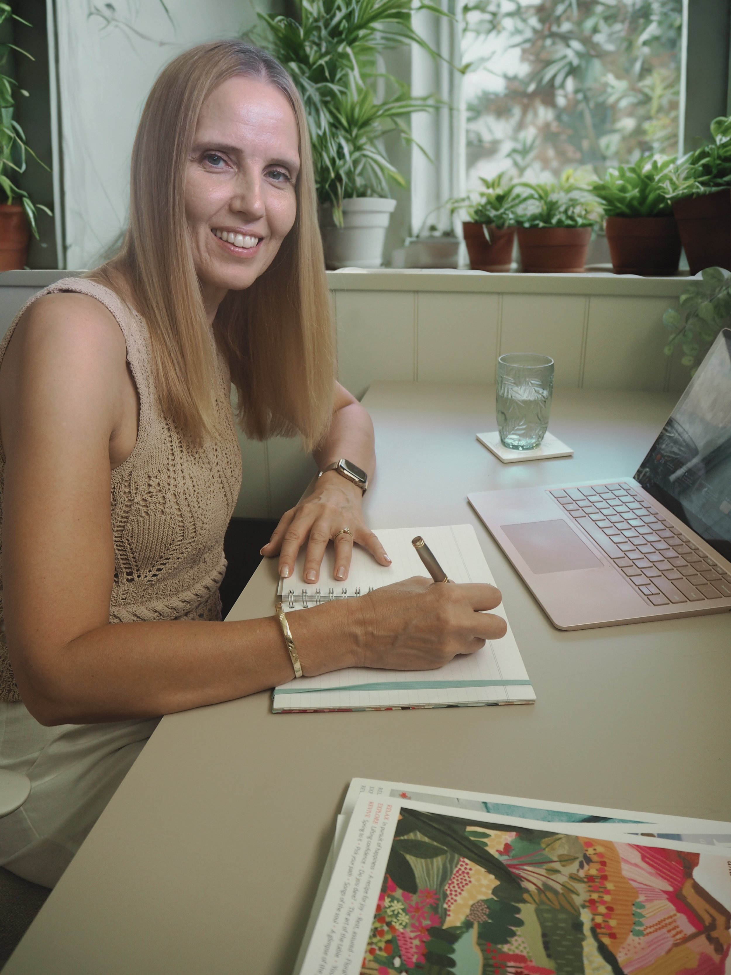 A woman with long blonde hair writing in a notebook at a desk, surrounded by green potted plants, with a laptop and a glass of water nearby.