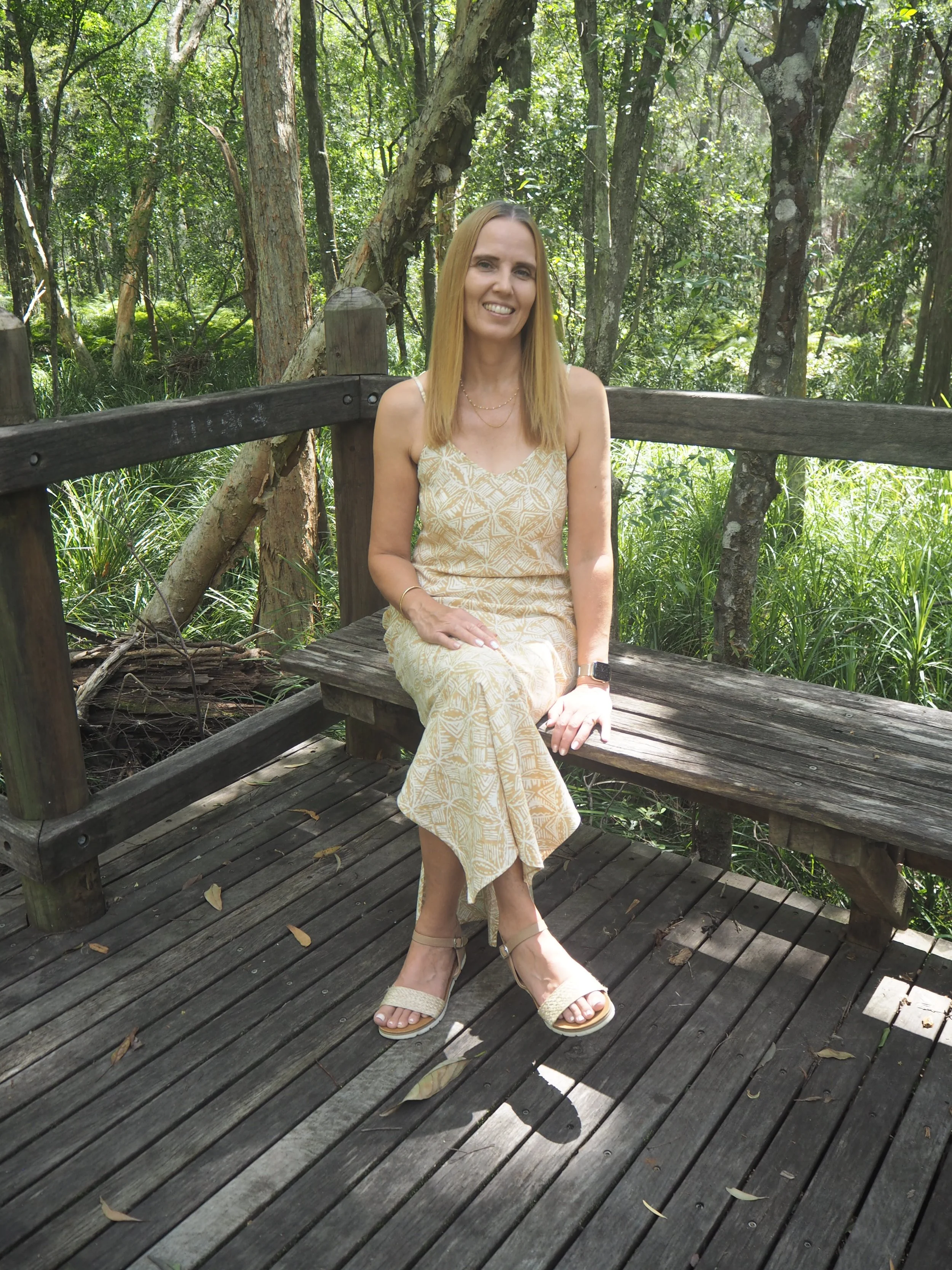 Woman in a light-colored dress sitting on a wooden bench in a forested area with trees and green foliage.
