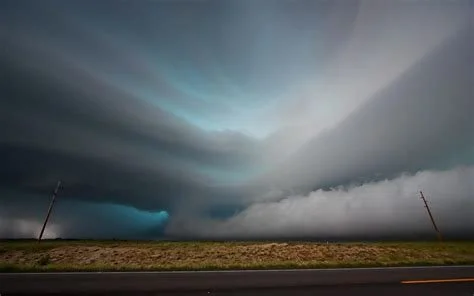 A dramatic storm cloud formation over a rural landscape with power poles and a highway.